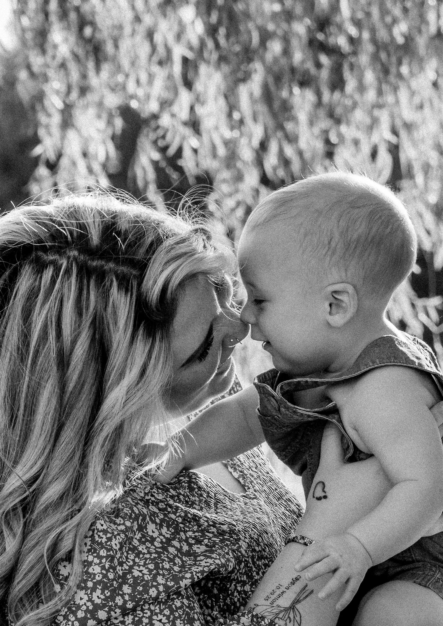 Black and white photo of a mother tenderly touching noses with her smiling baby outdoors under a willow tree.