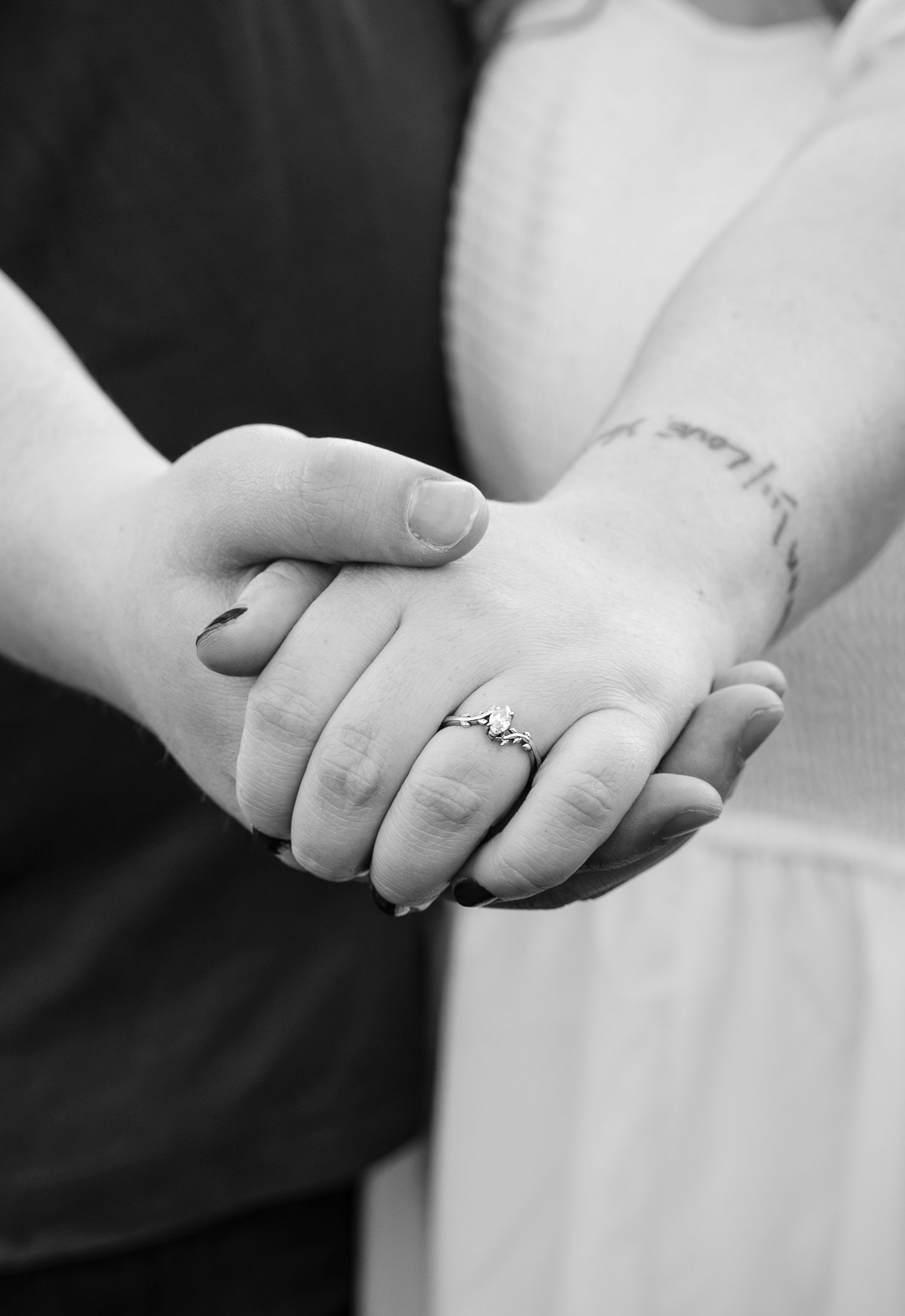 Black and white close-up of couple holding hands showing engagement ring and wrist tattoo detail.