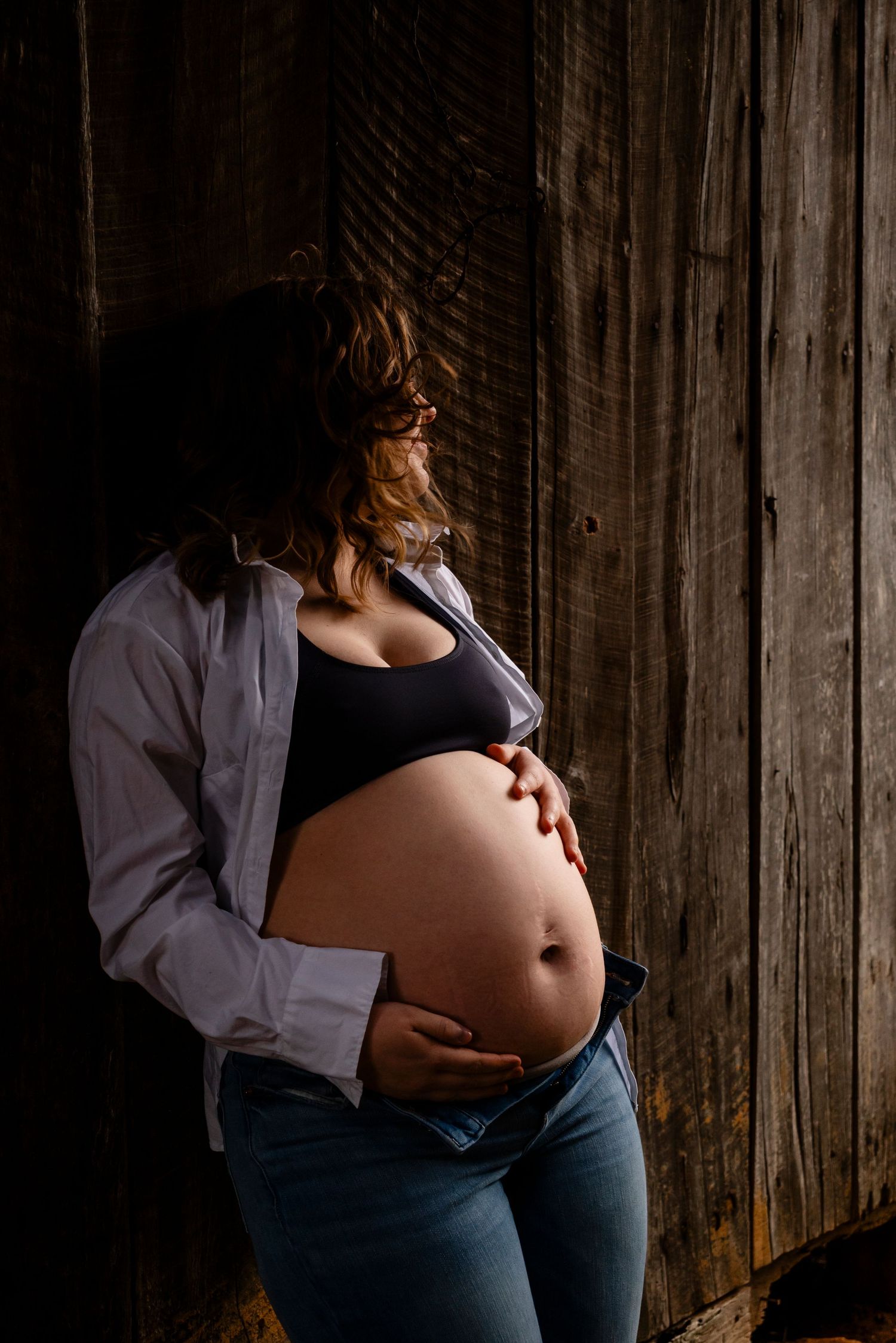Pregnant woman in white shirt and jeans holds baby bump while leaning against dark wooden barn wall, dramatic lighting.