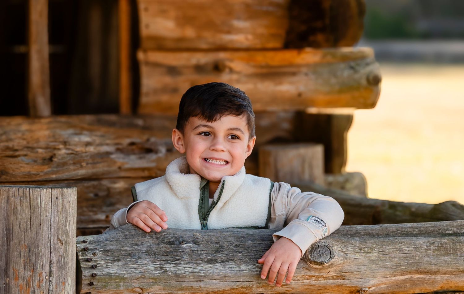 Adorable young boy peeks over a weathered wooden fence rail beside a rustic log cabin outdoors.
