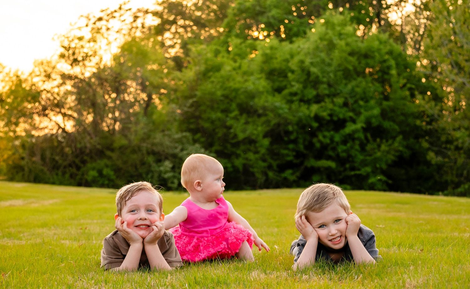 Two boys and a baby girl in pink dress pose lying on lush green grass at sunset.