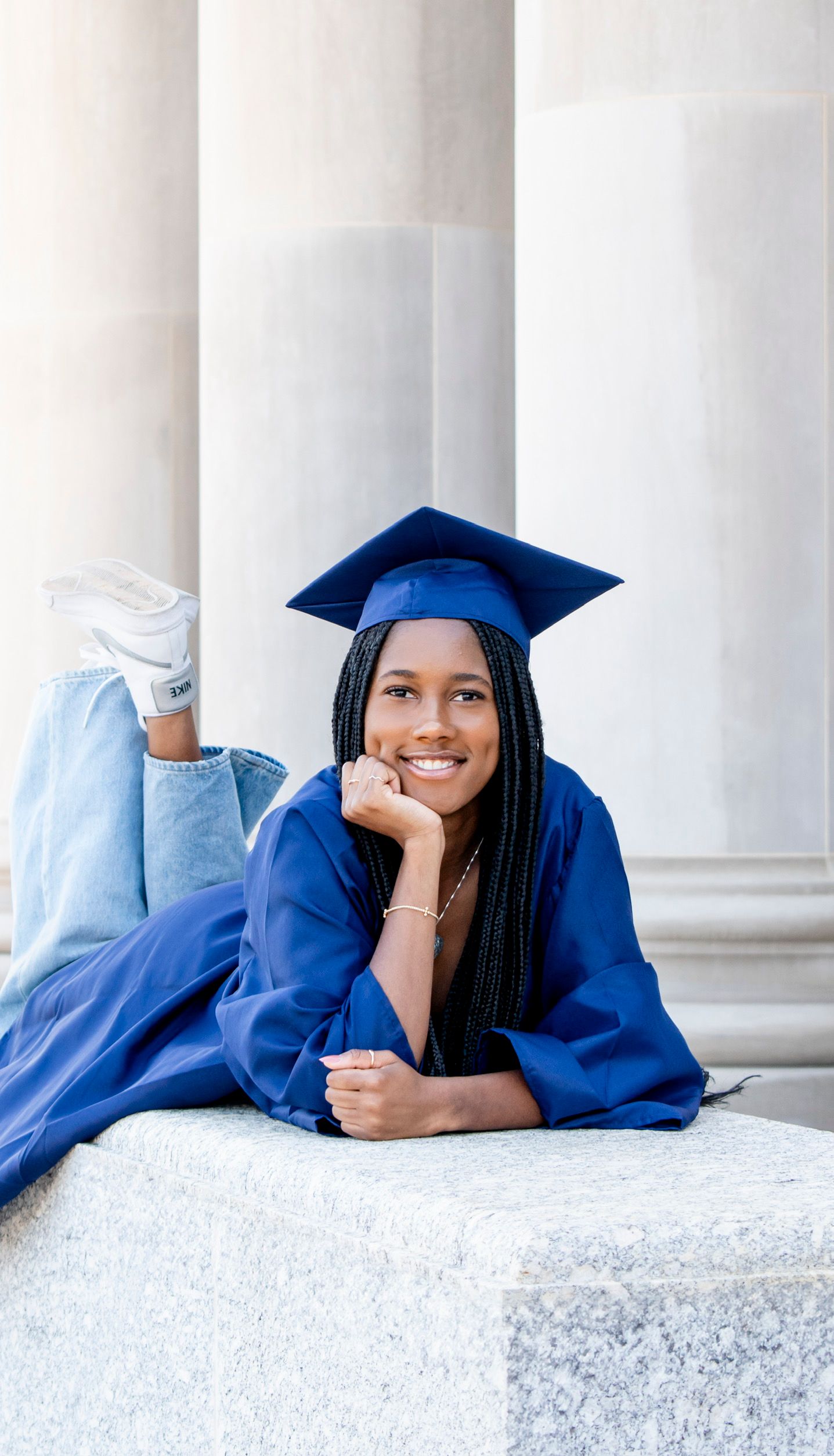 Smiling graduate in blue cap and gown lying on stone steps, wearing jeans and white sneakers.