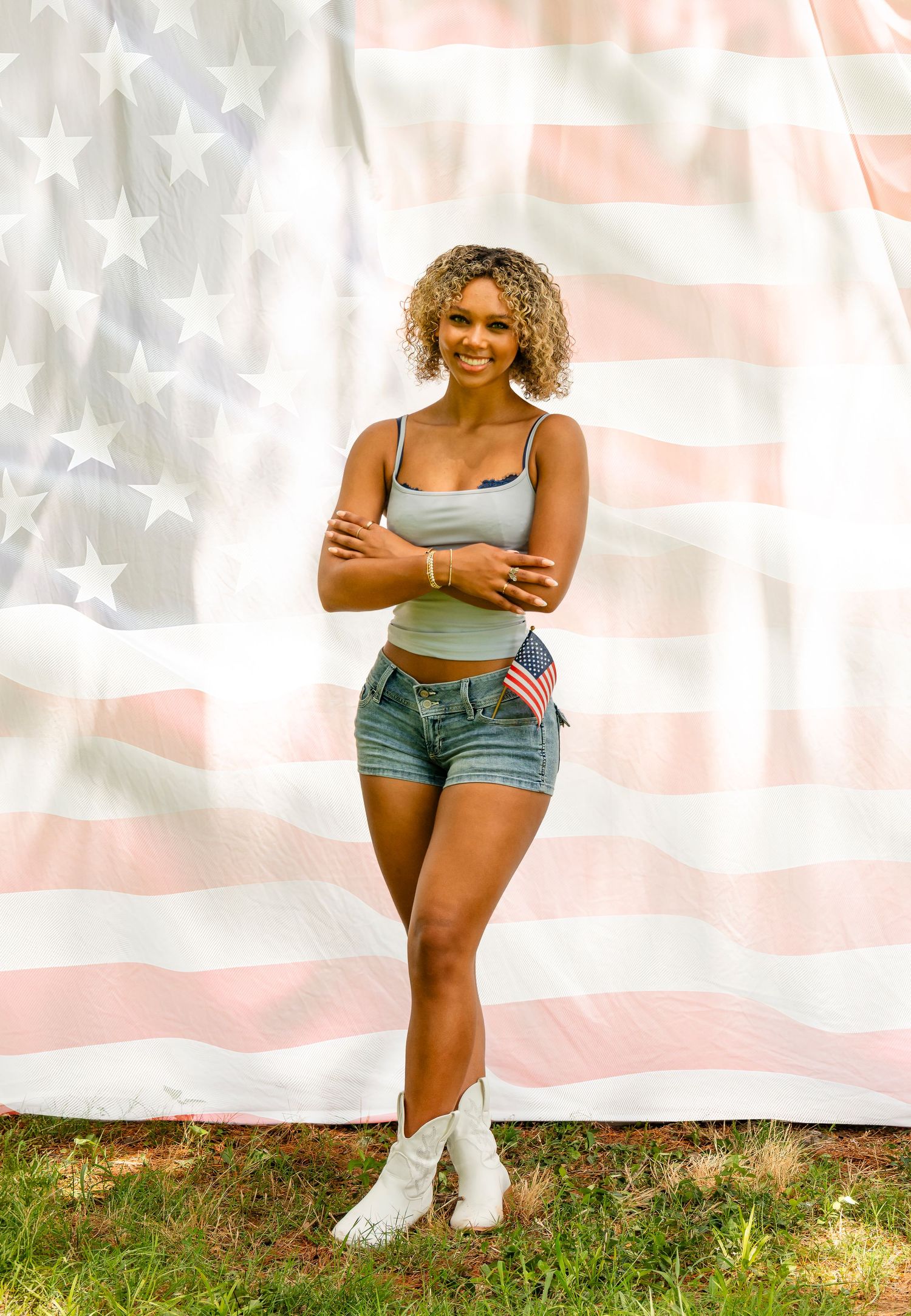Smiling woman with curly hair stands before a large American flag wearing denim shorts and white boots.