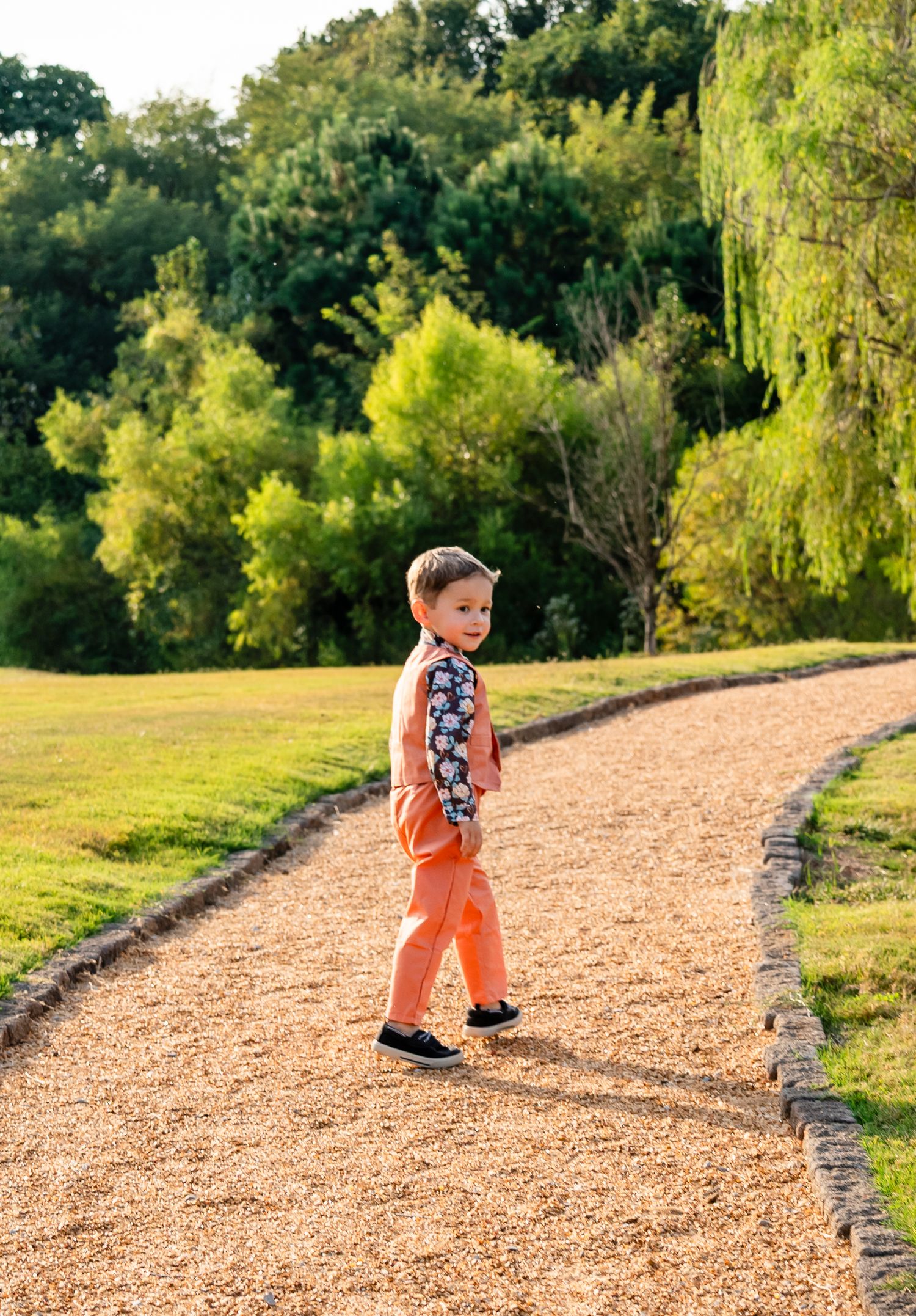 Young boy in orange pants and floral shirt walking on a gravel path surrounded by lush green trees.