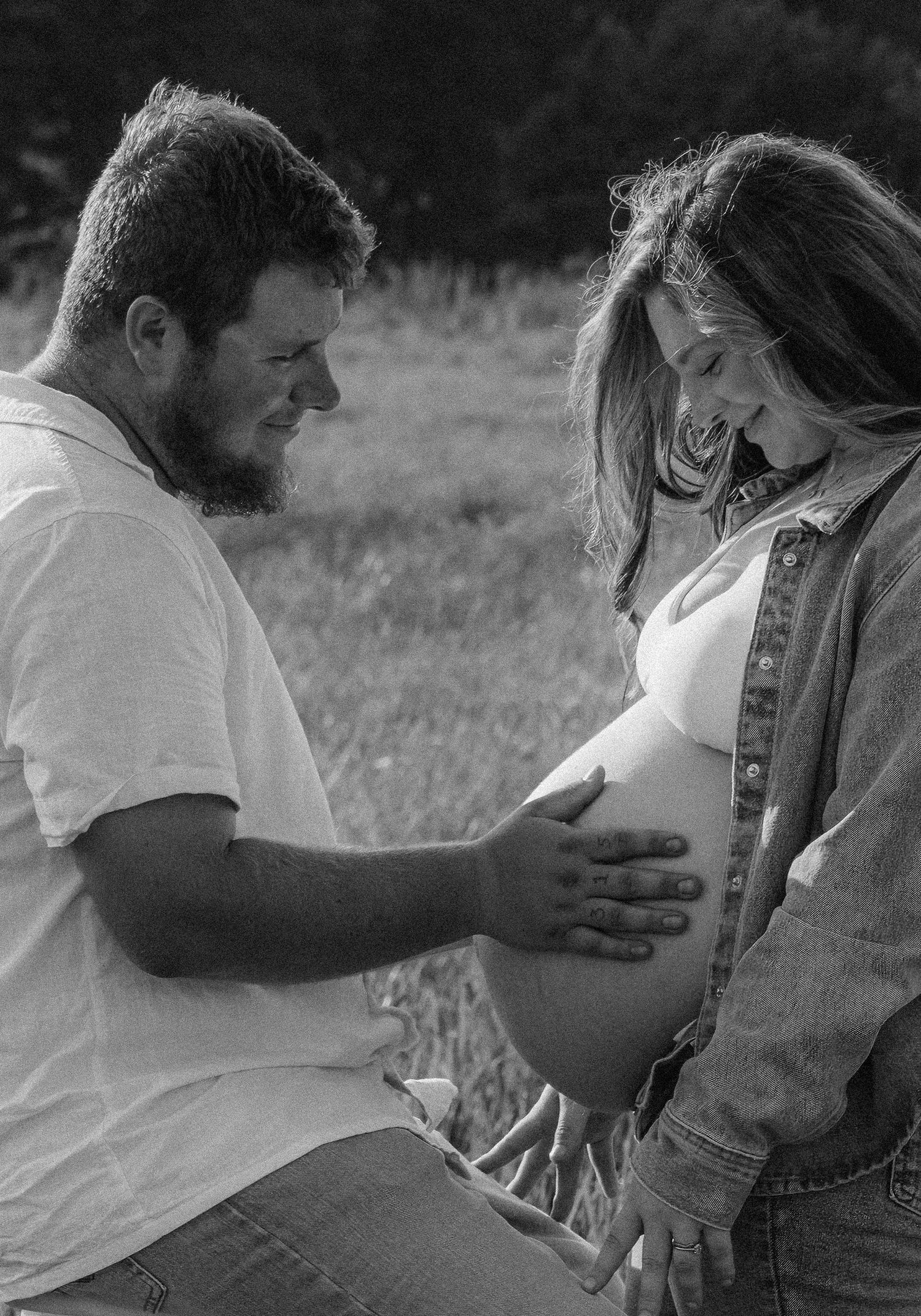 Black and white maternity photo of a couple smiling at pregnant belly outdoors in a field.