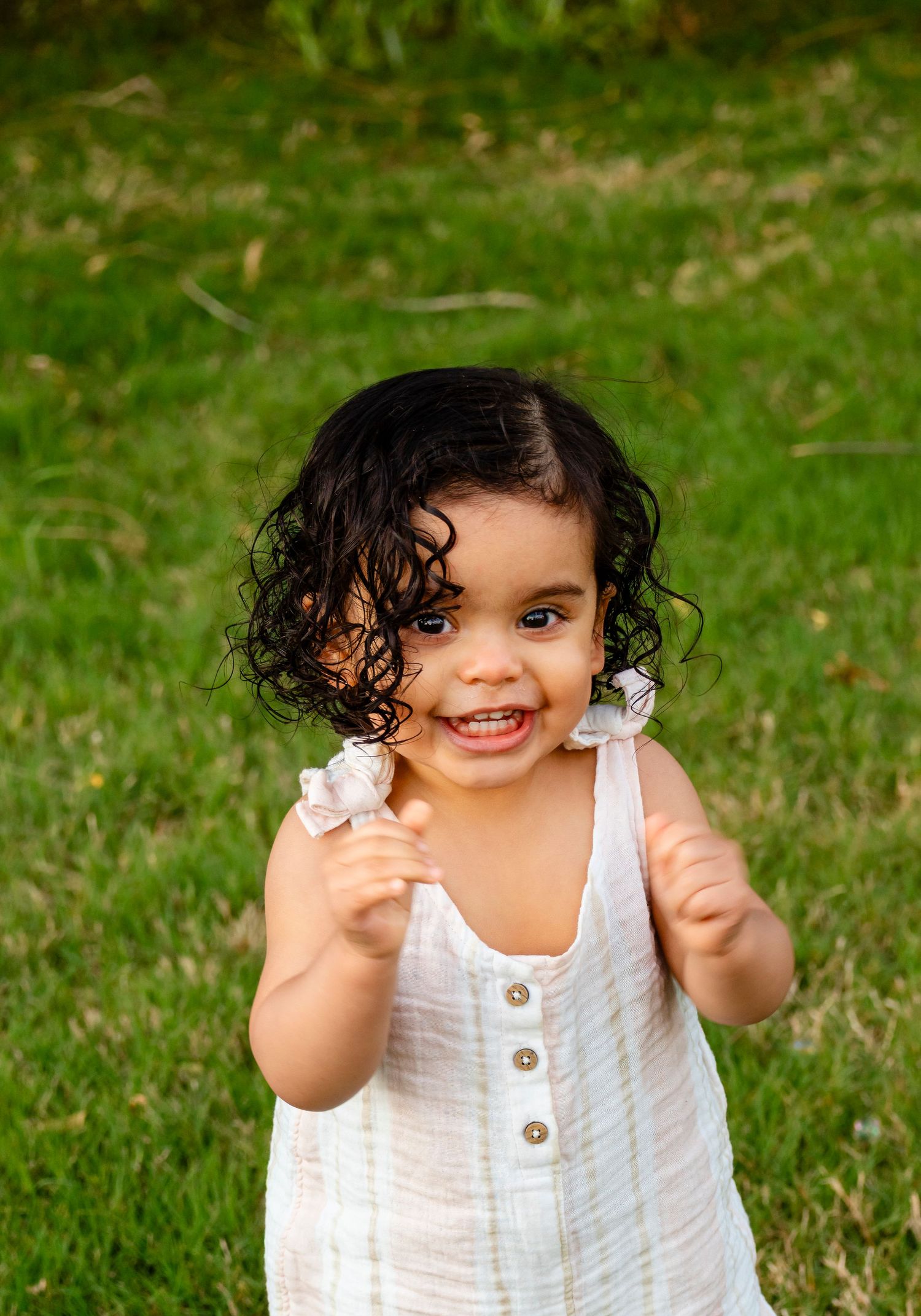 Smiling toddler girl with curly dark hair wearing a white dress, standing on green grass outdoors.