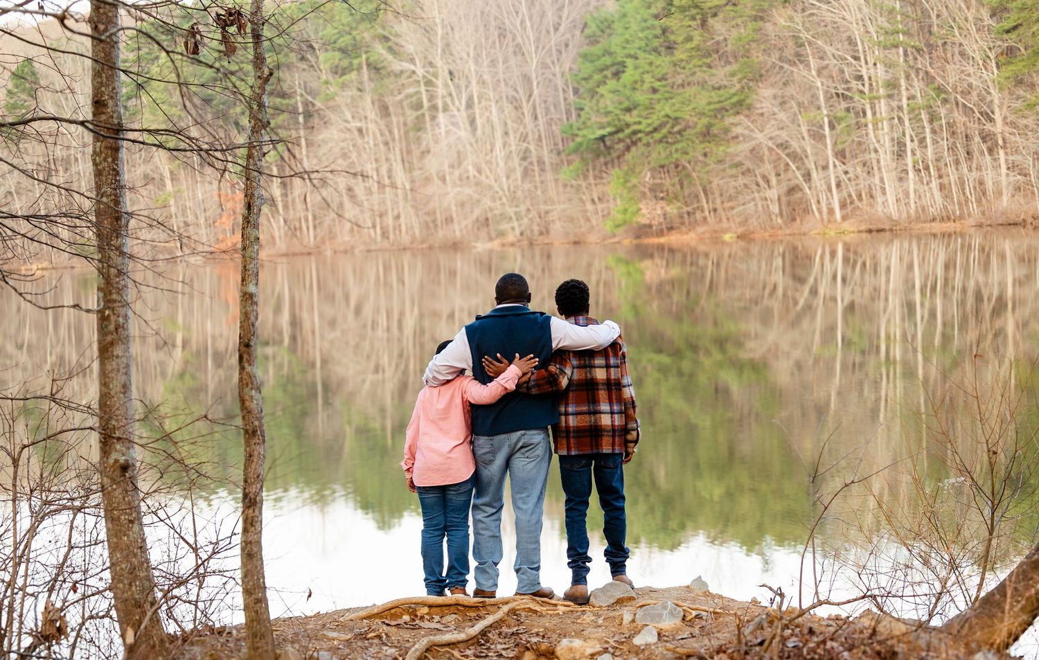 Three children stand arm-in-arm on a rocky overlook gazing at a serene forest lake in autumn.