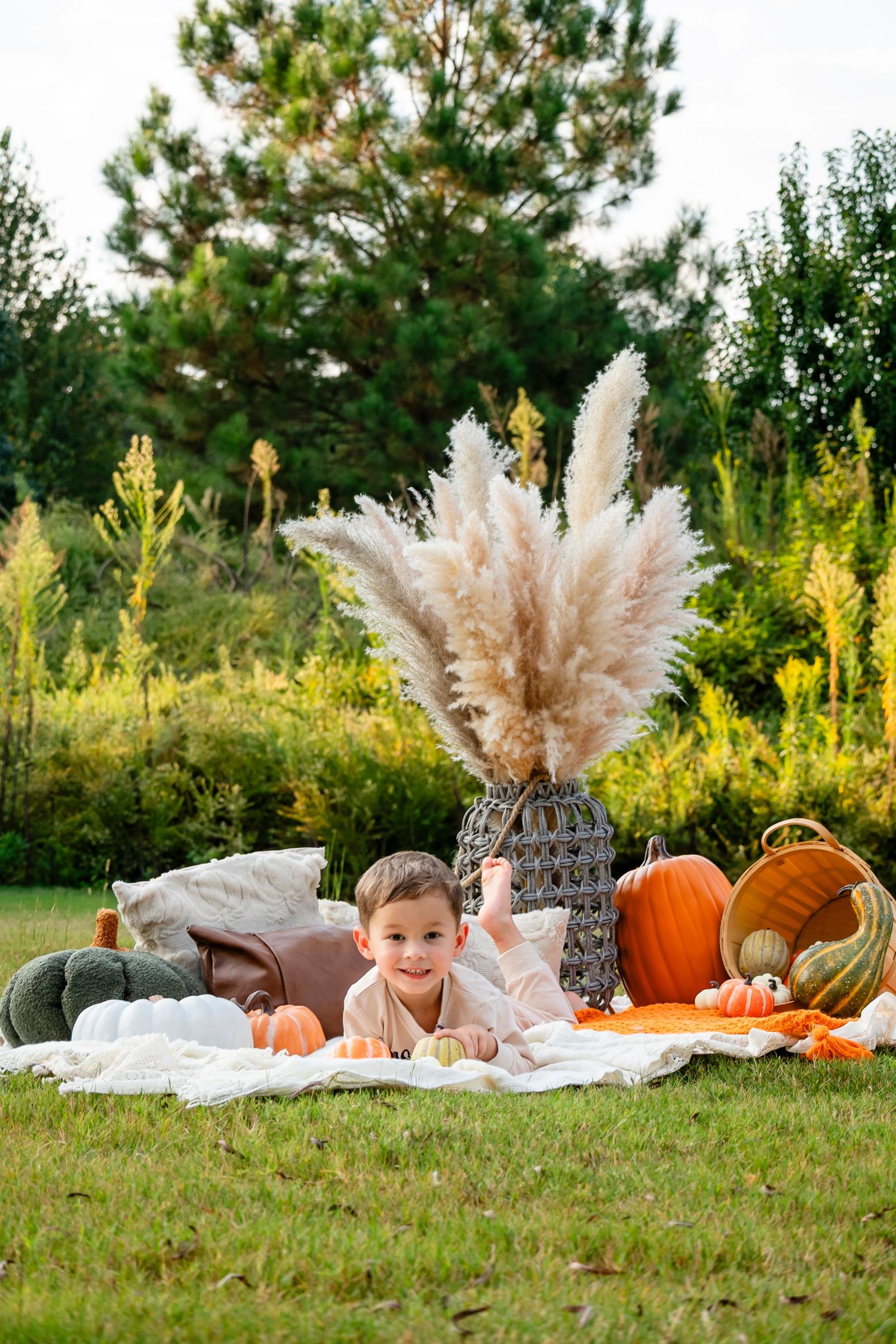Young boy lying on white blanket surrounded by pumpkins and pampas grass in autumn garden.