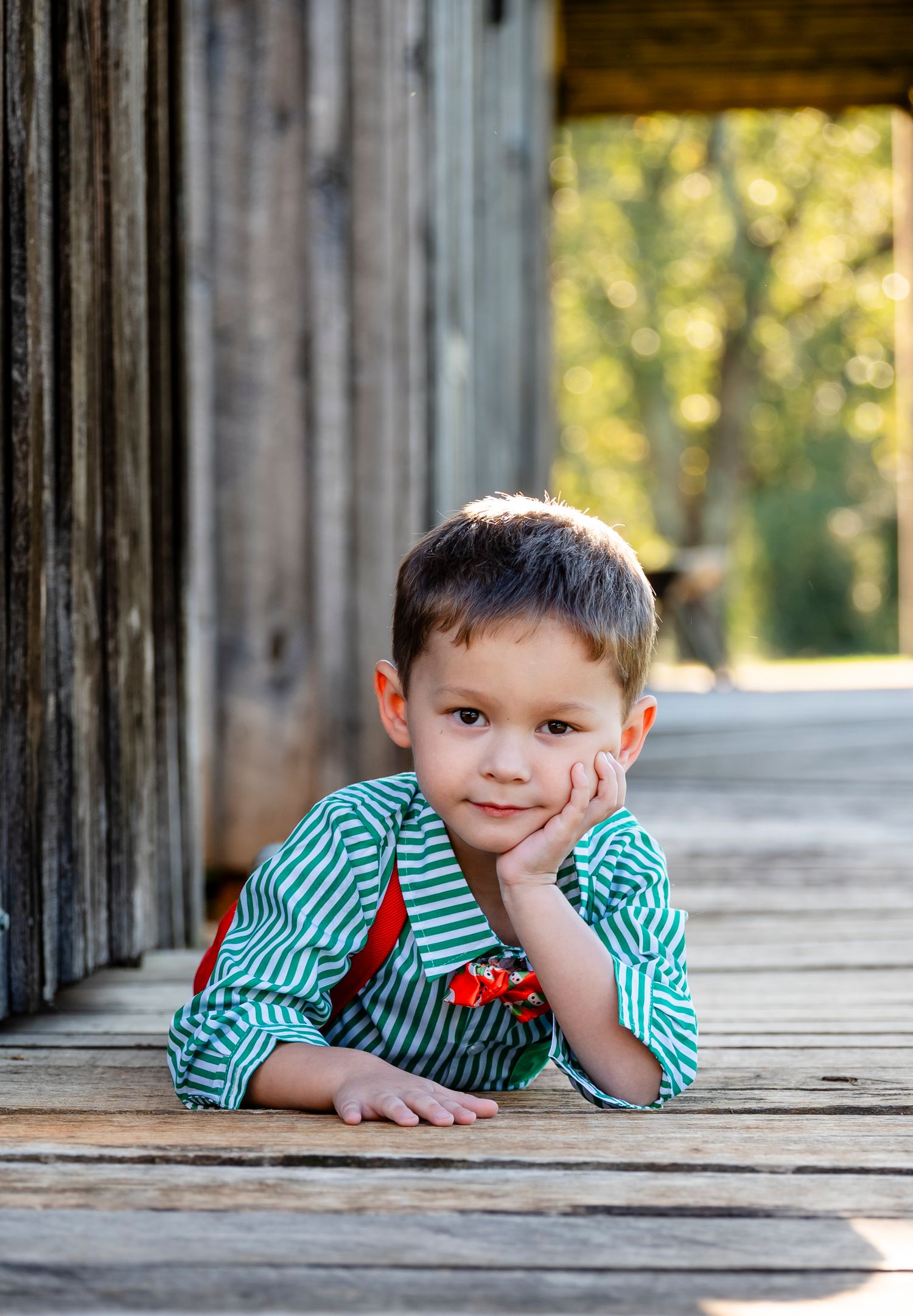 Young boy in green striped shirt lying on wooden deck with chin resting on hand, looking at camera.