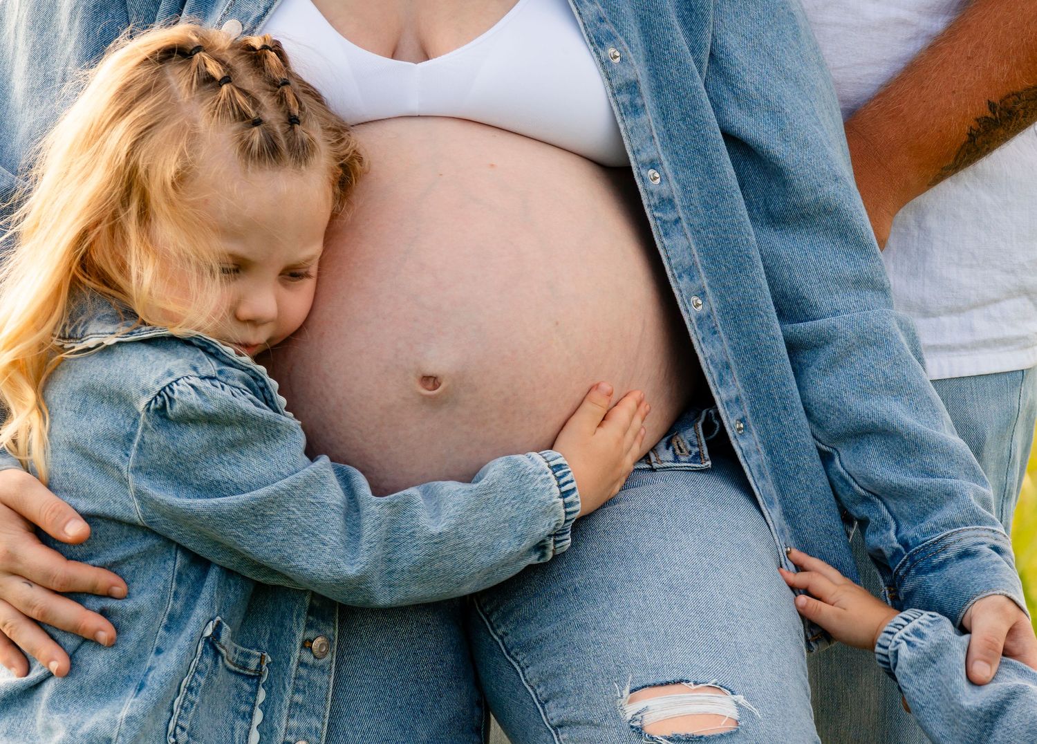 A young girl hugging her pregnant mother's bare belly outdoors, wearing matching denim outfits.