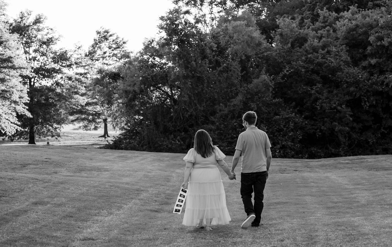 Black and white photo of a couple holding hands walking through a lush green park in wedding attire.