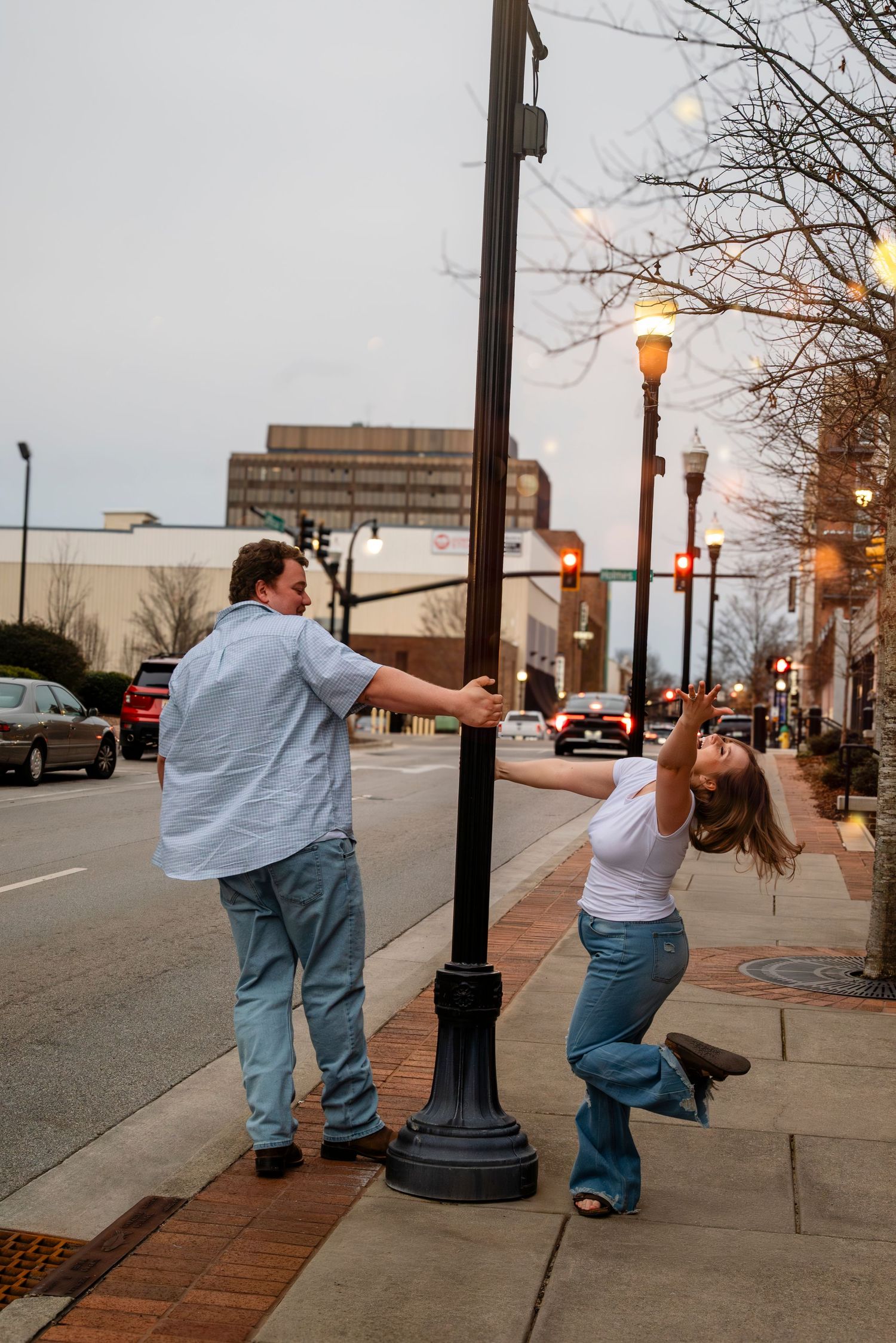 Couple playfully spinning around a street lamp post on a city sidewalk at dusk.