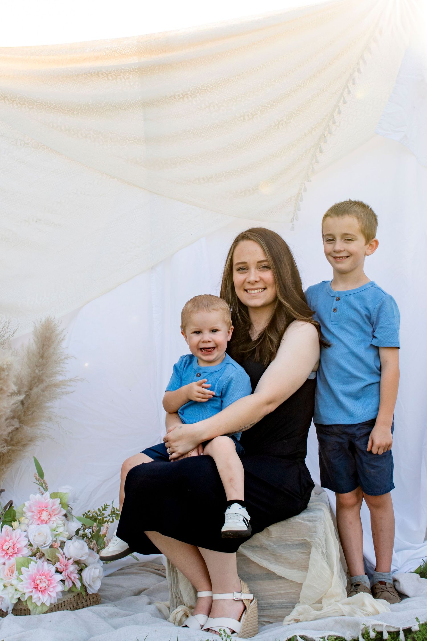 A smiling mother in a black dress holds a laughing toddler while an older boy in blue stands beside them outdoors.