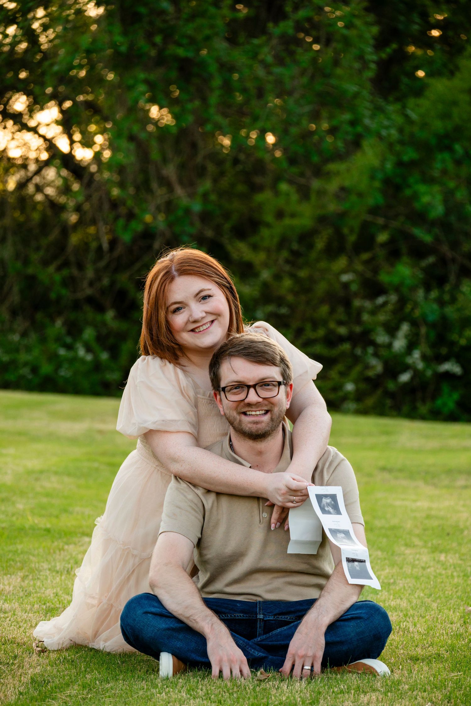 Happy couple sitting on grass holding ultrasound photos during outdoor pregnancy announcement photo session at sunset.