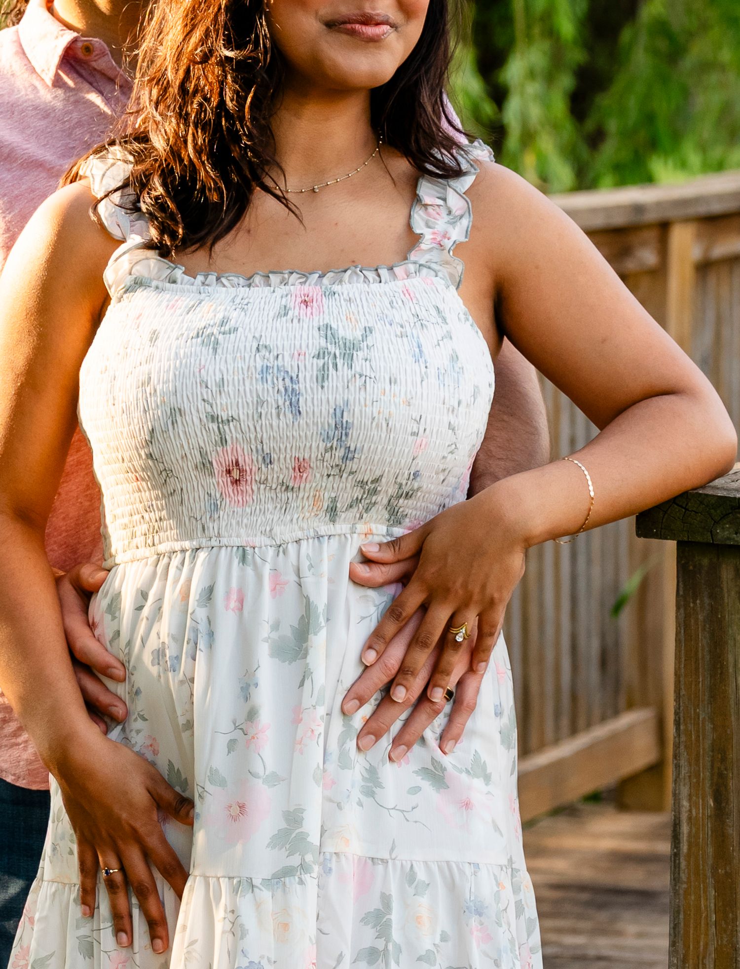 Pregnant woman in white floral smocked dress cradling baby bump outdoors on wooden bridge.