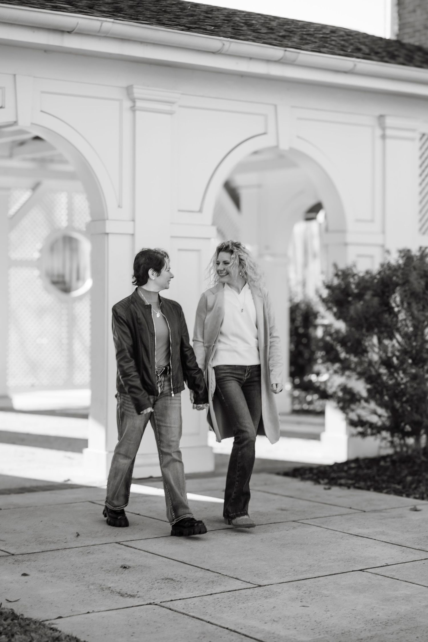 Black and white photo of two women walking and talking on a sidewalk in front of a white arched building.