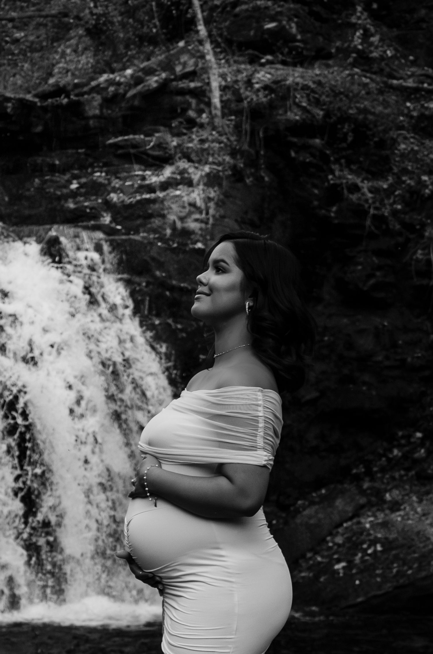 Black and white maternity photo of a smiling pregnant woman in a white off-shoulder dress posing before a waterfall.