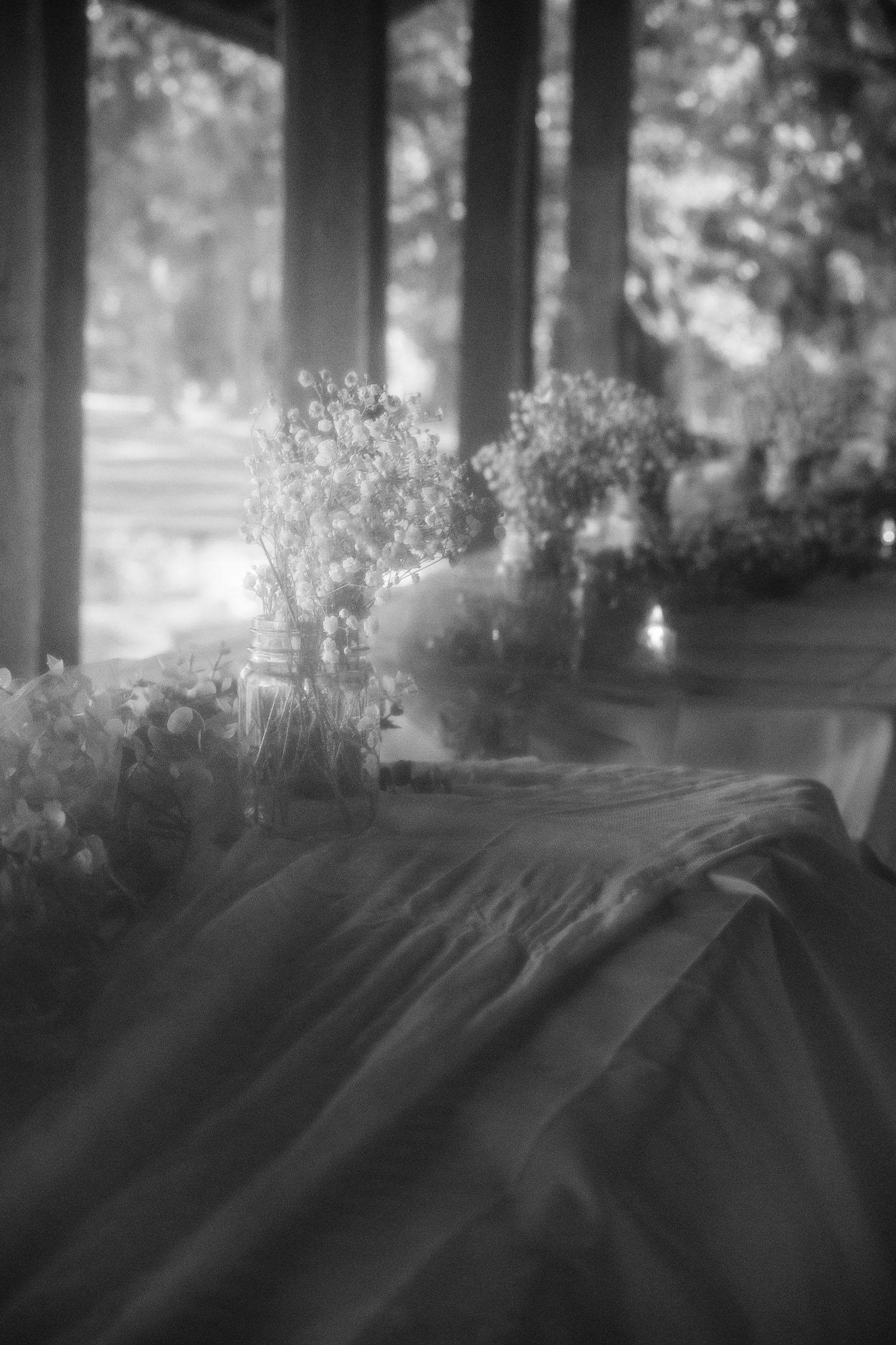 Black and white photo of floral centerpieces on a draped table near large windows with soft natural light.