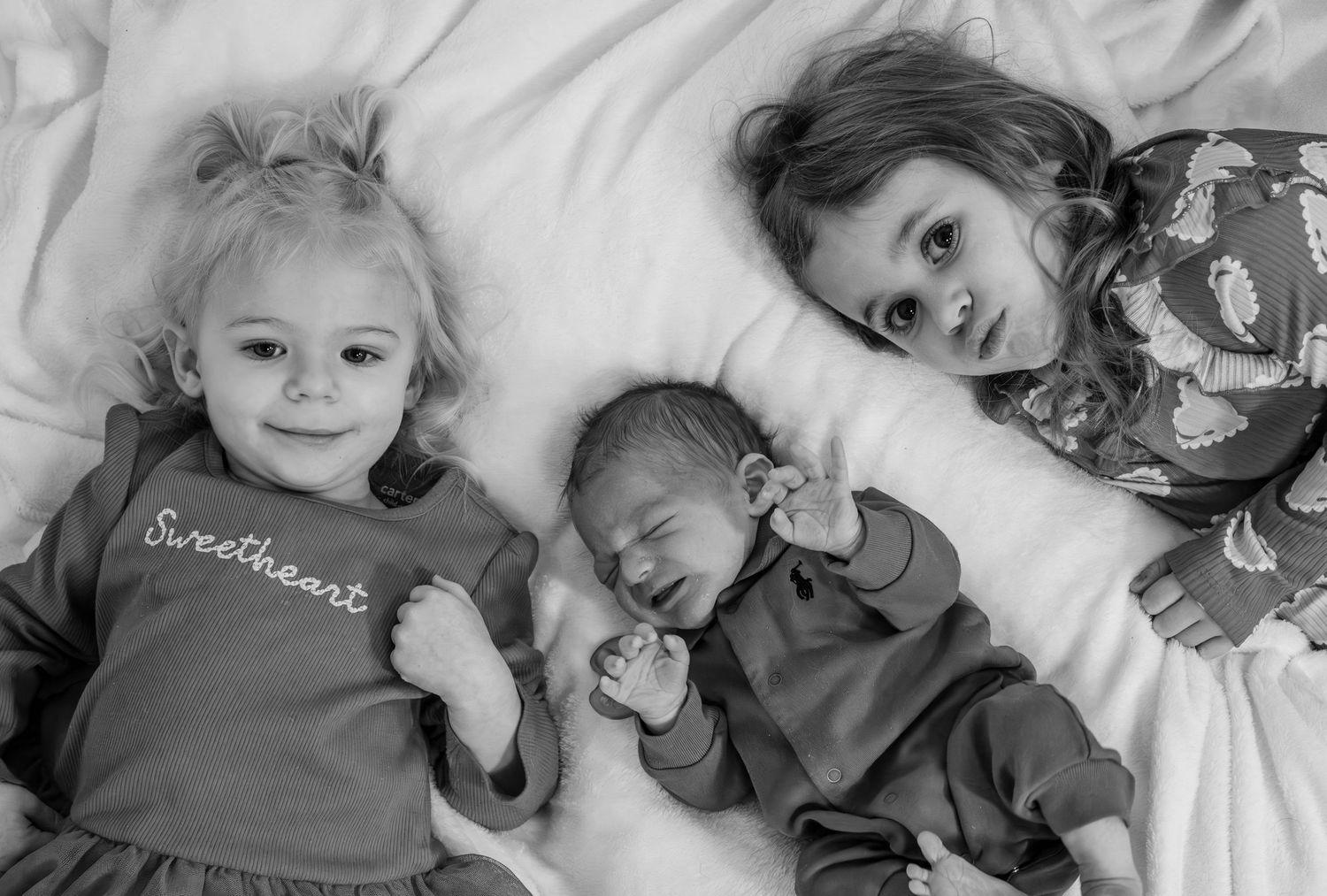 Black and white photo of two toddler girls and a newborn baby lying together on a white bed.