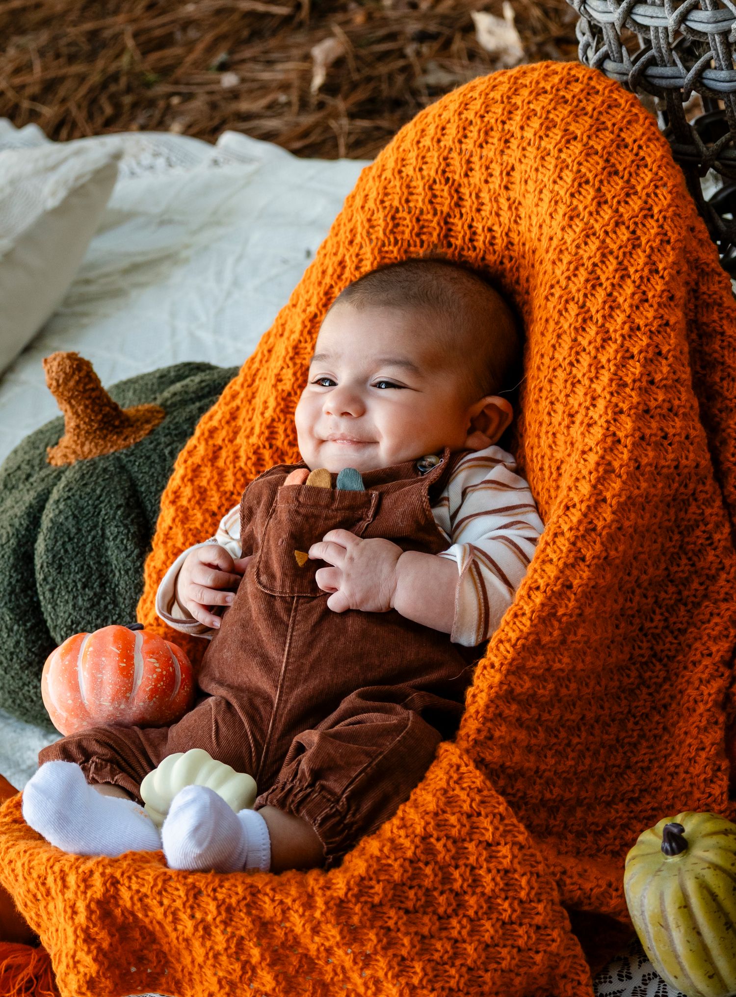 Happy baby smiling in orange knit blanket surrounded by decorative pumpkins in fall setting.