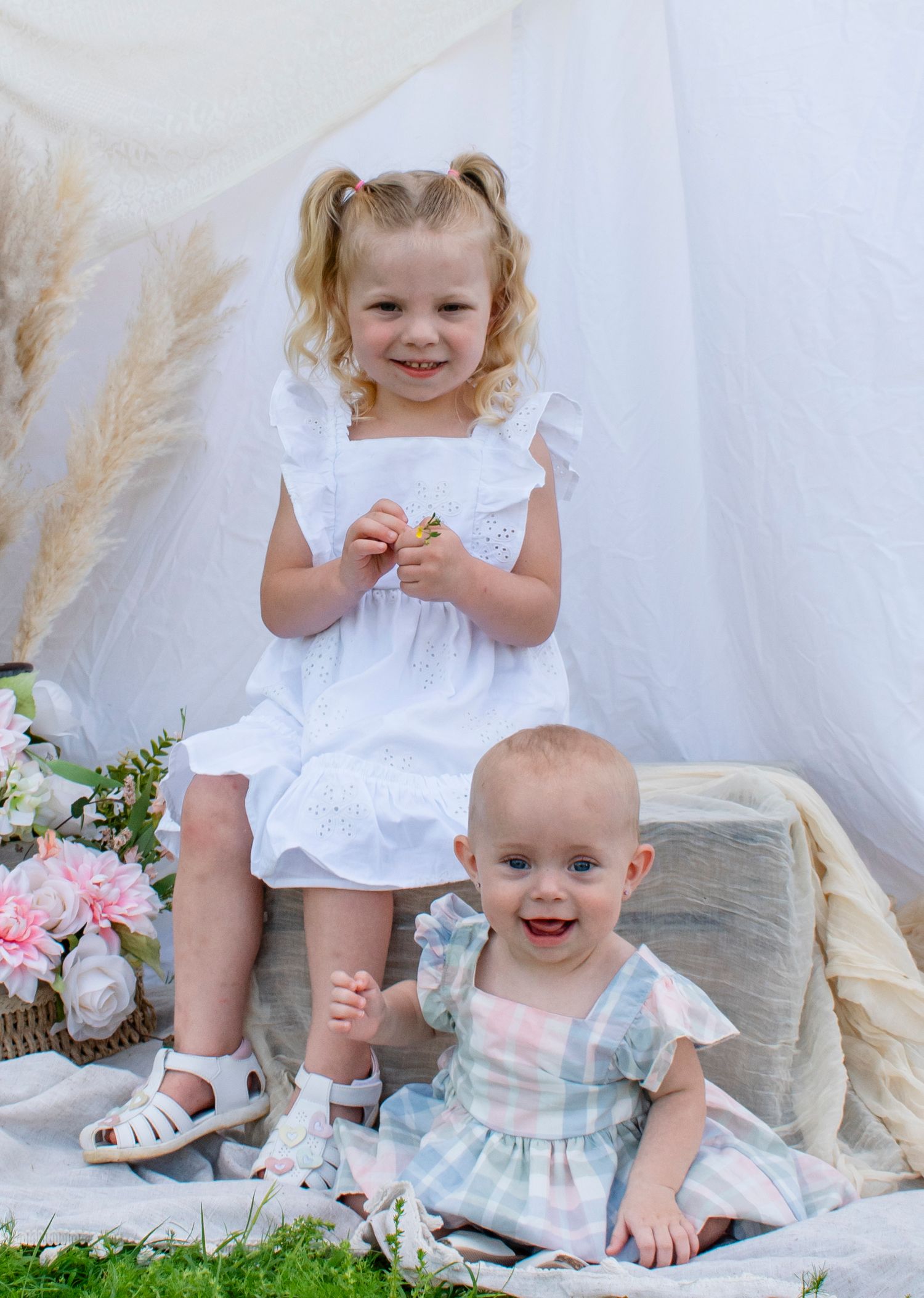 Two young sisters pose together, an older curly-haired toddler in a white dress standing beside her smiling baby sister.
