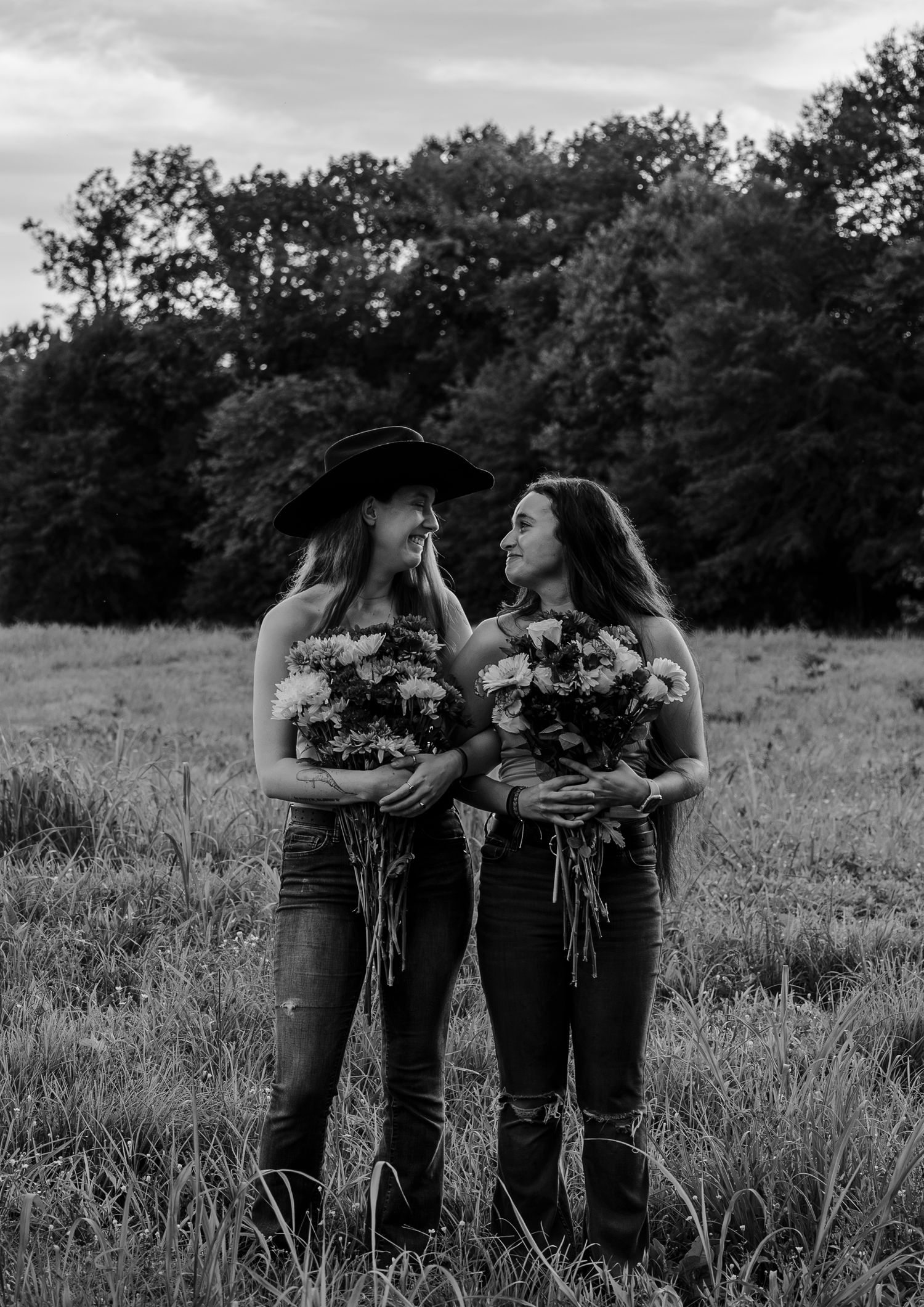 Black and white photo of two women laughing together in a field, holding flower bouquets, one in a cowboy hat.