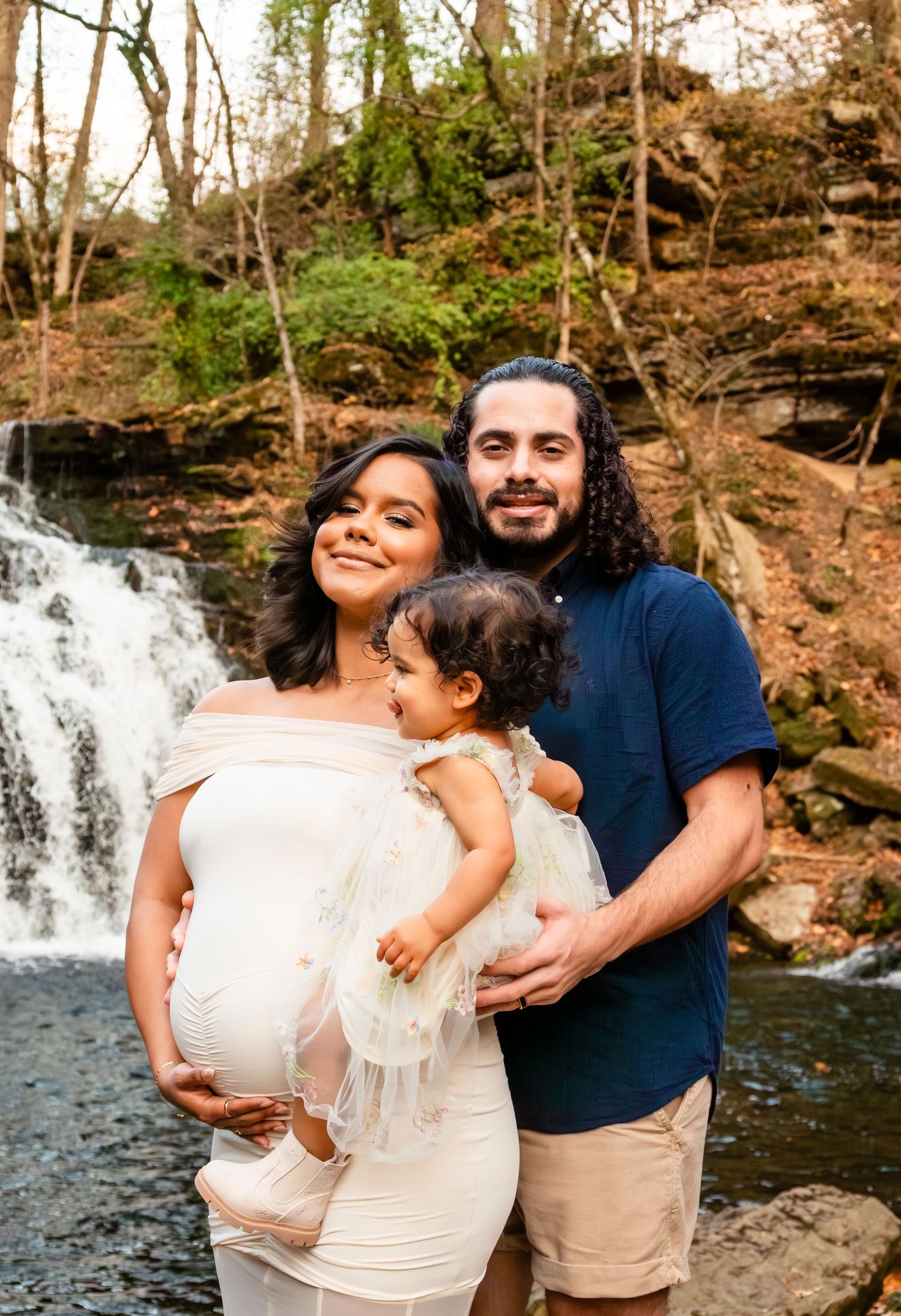 Pregnant woman in white dress with partner and toddler posing before a waterfall in a lush forest setting.