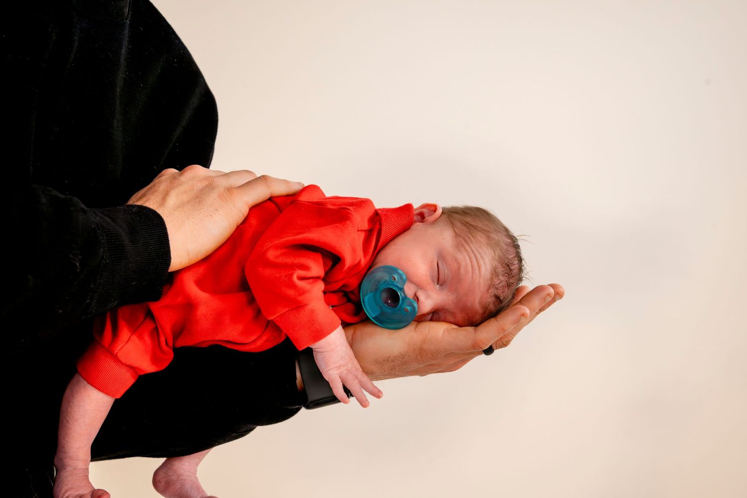 Newborn baby in red outfit with a blue pacifier resting on one hand against white background.