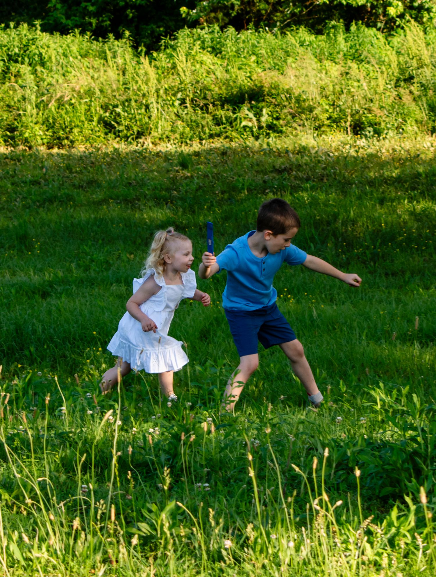 Two young children playing in a sunny green meadow, girl in white dress chasing boy in blue shirt.