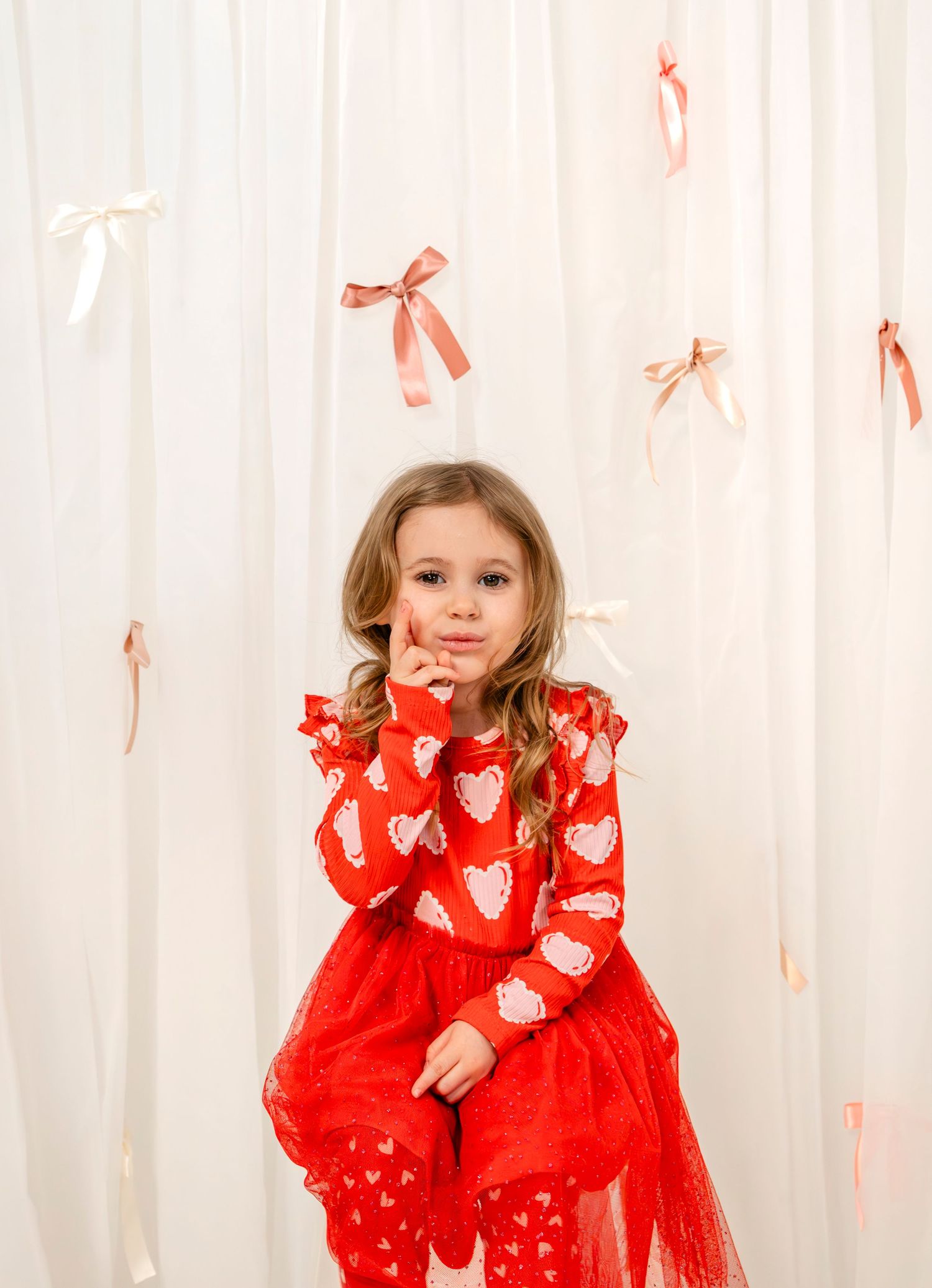 Young girl in a red heart-print dress and tutu poses thoughtfully in front of a white bow backdrop.