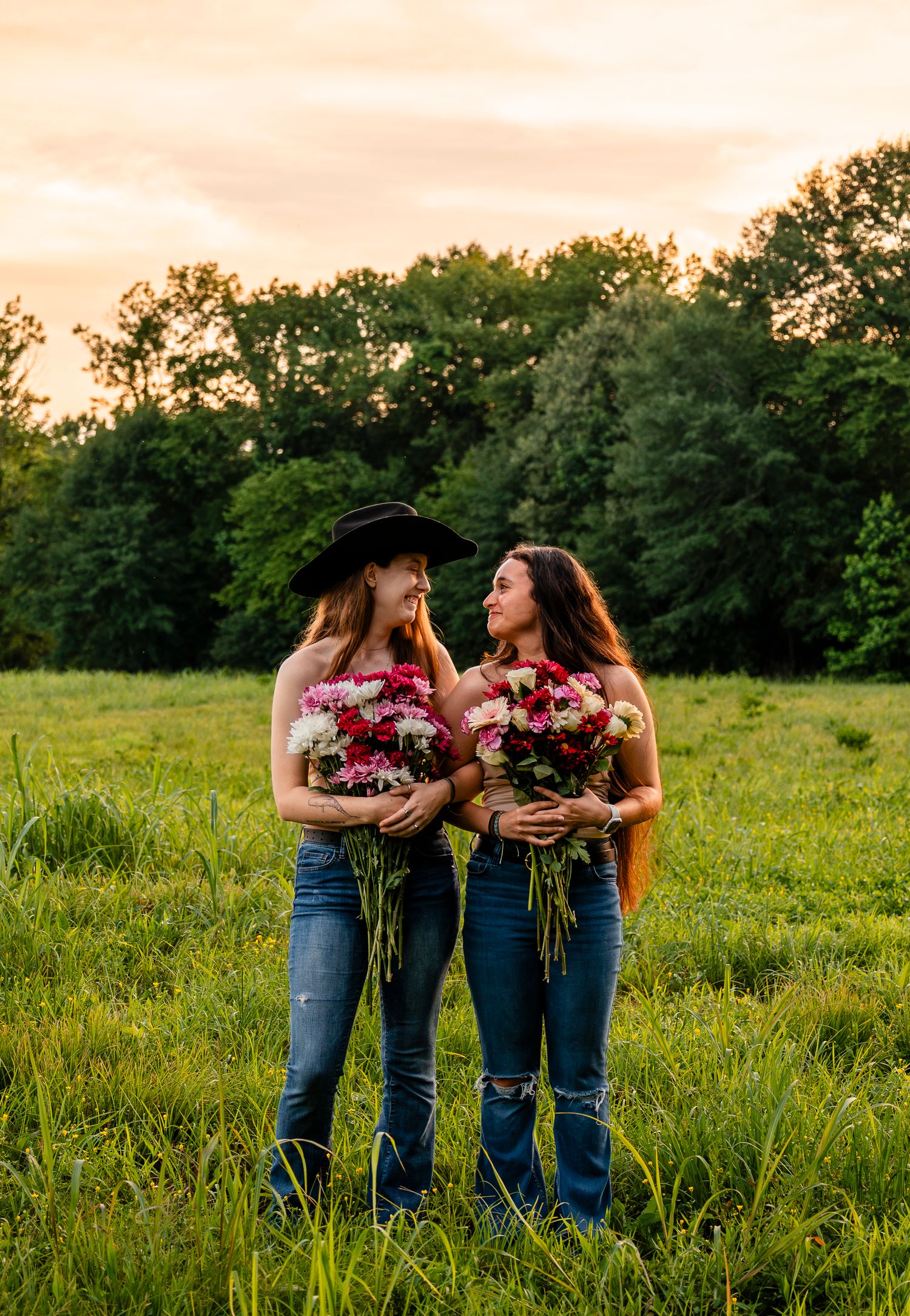 Two smiling women in jeans hold vibrant flower bouquets while standing together in a green field at golden hour.
