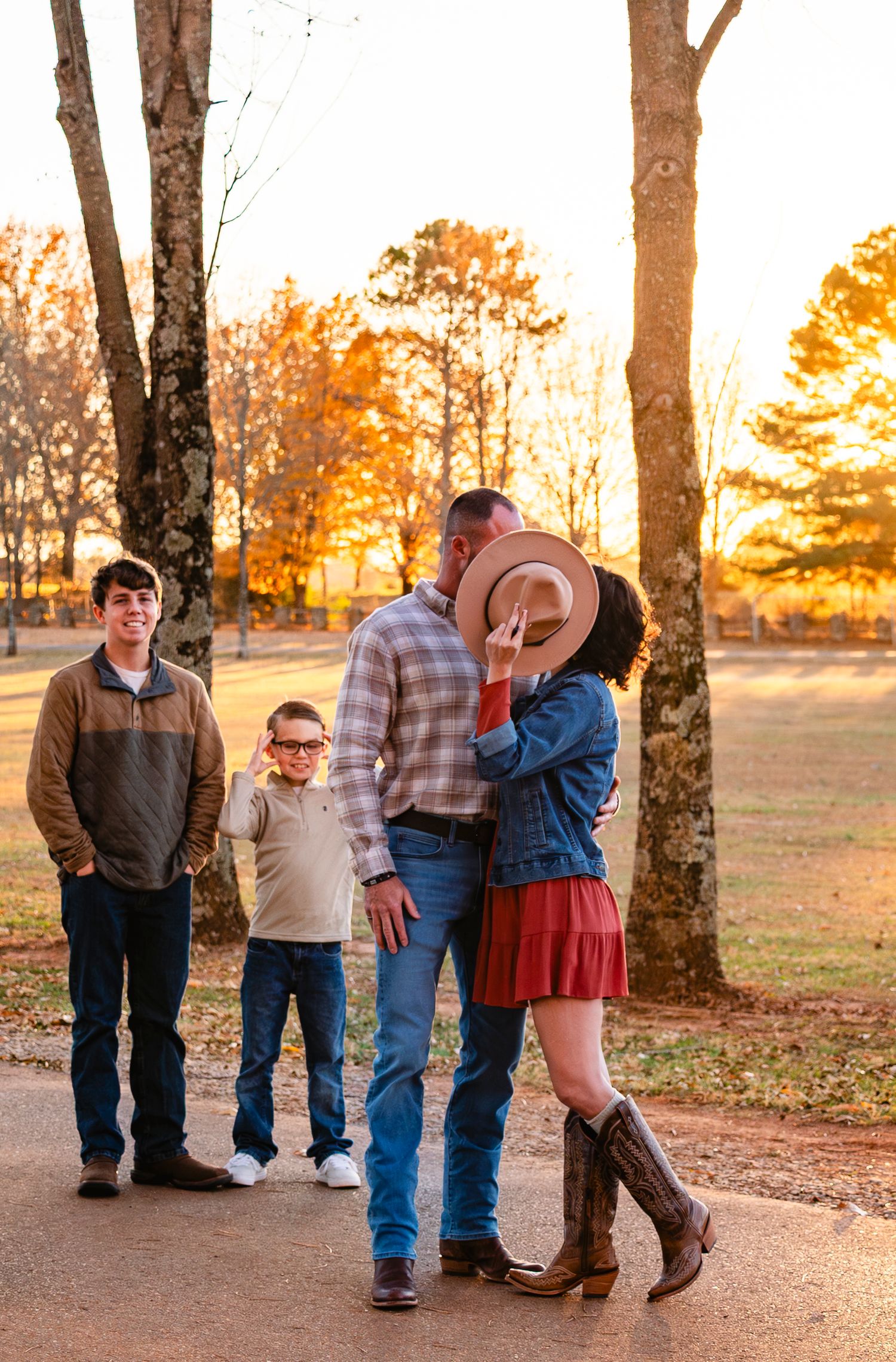 Couple shares a kiss hidden behind a cowboy hat while two boys watch during a fall family photo session.