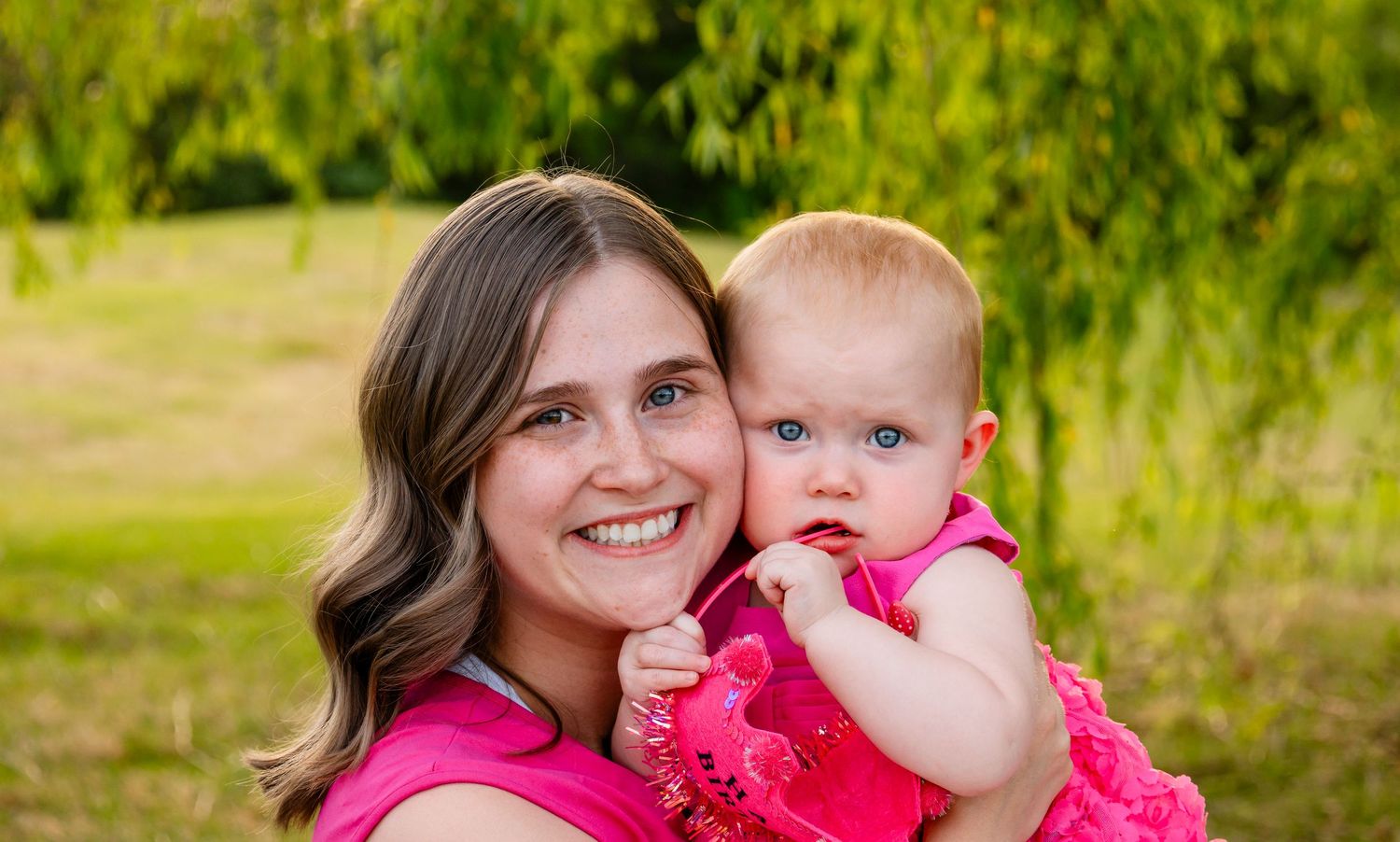 Smiling young woman in pink top holds adorable baby girl in matching pink dress outdoors.