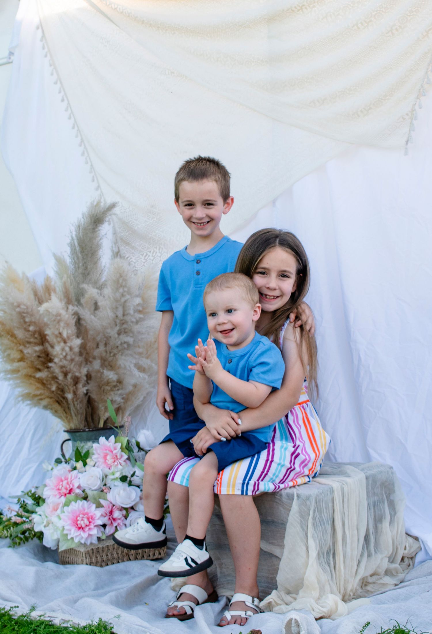 Three siblings pose together, two older children in blue outfits embracing a smiling toddler in a blue shirt.