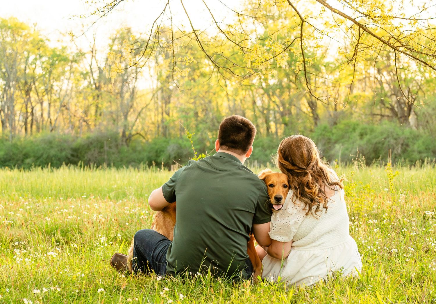 Couple and their golden dog sitting together in a flower-filled meadow surrounded by spring trees.
