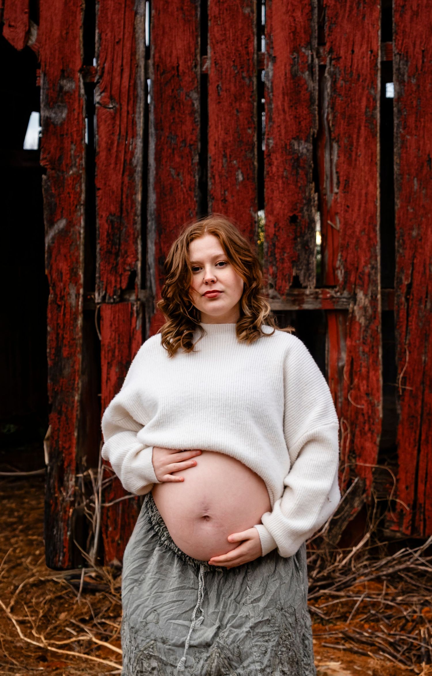 Pregnant woman in white sweater and gray skirt cradling baby bump in front of rustic red barn wall.