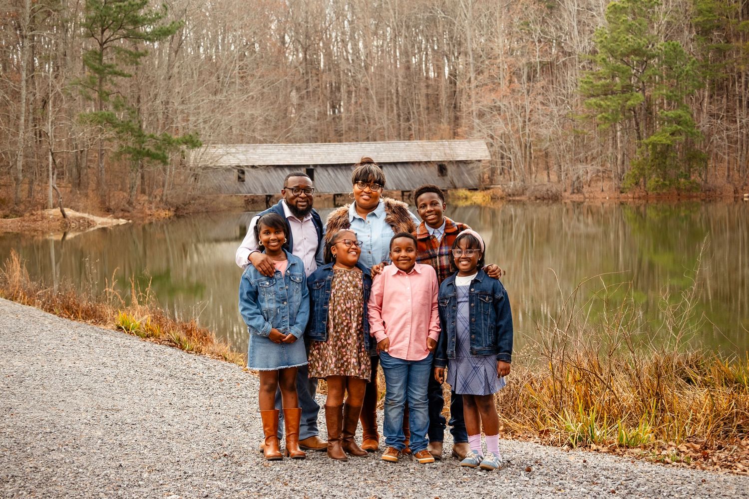 Large family group smiling together on a gravel path by a pond in bare winter woods.