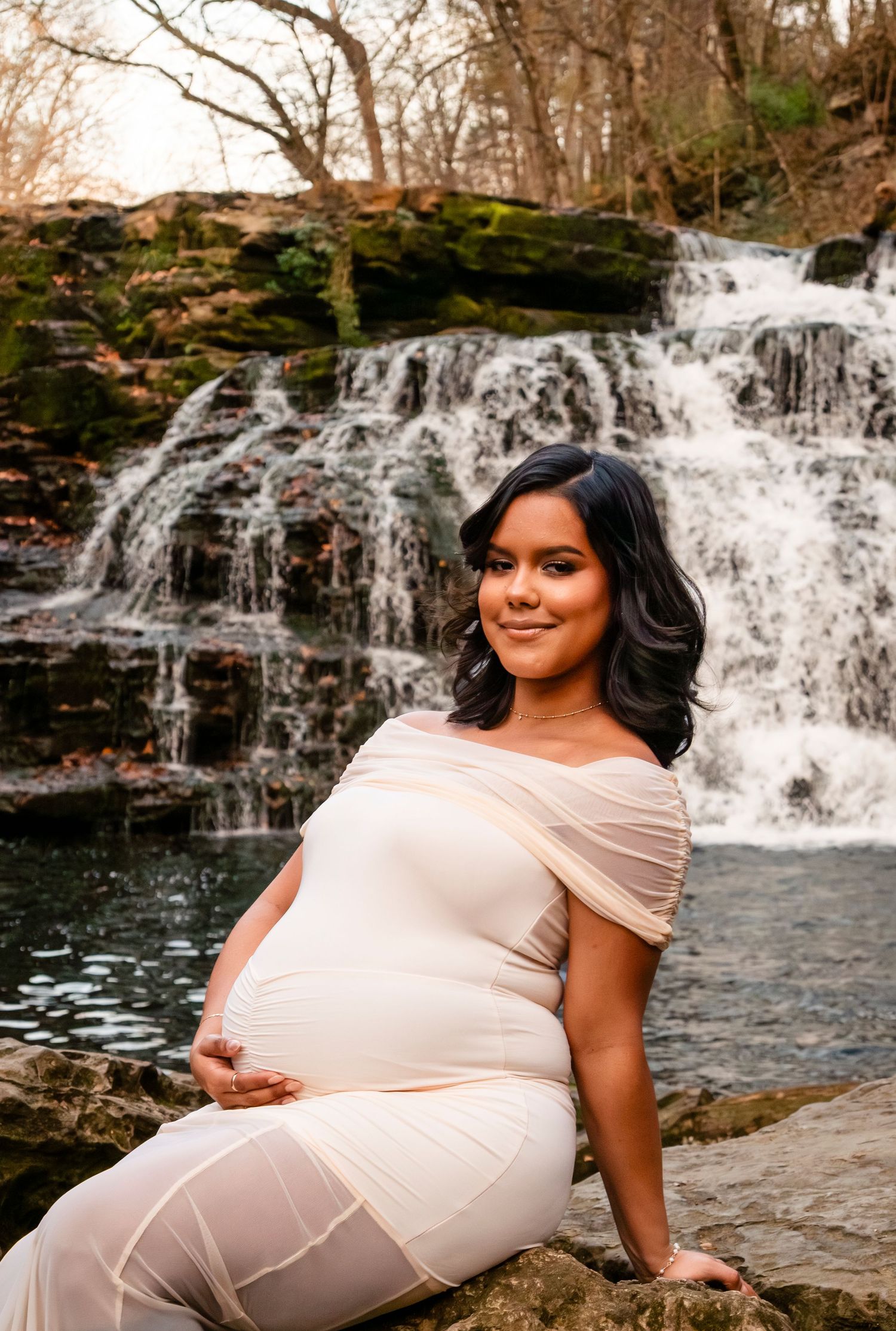 Glowing pregnant woman in cream off-shoulder gown sits on rocks before a picturesque cascading waterfall.