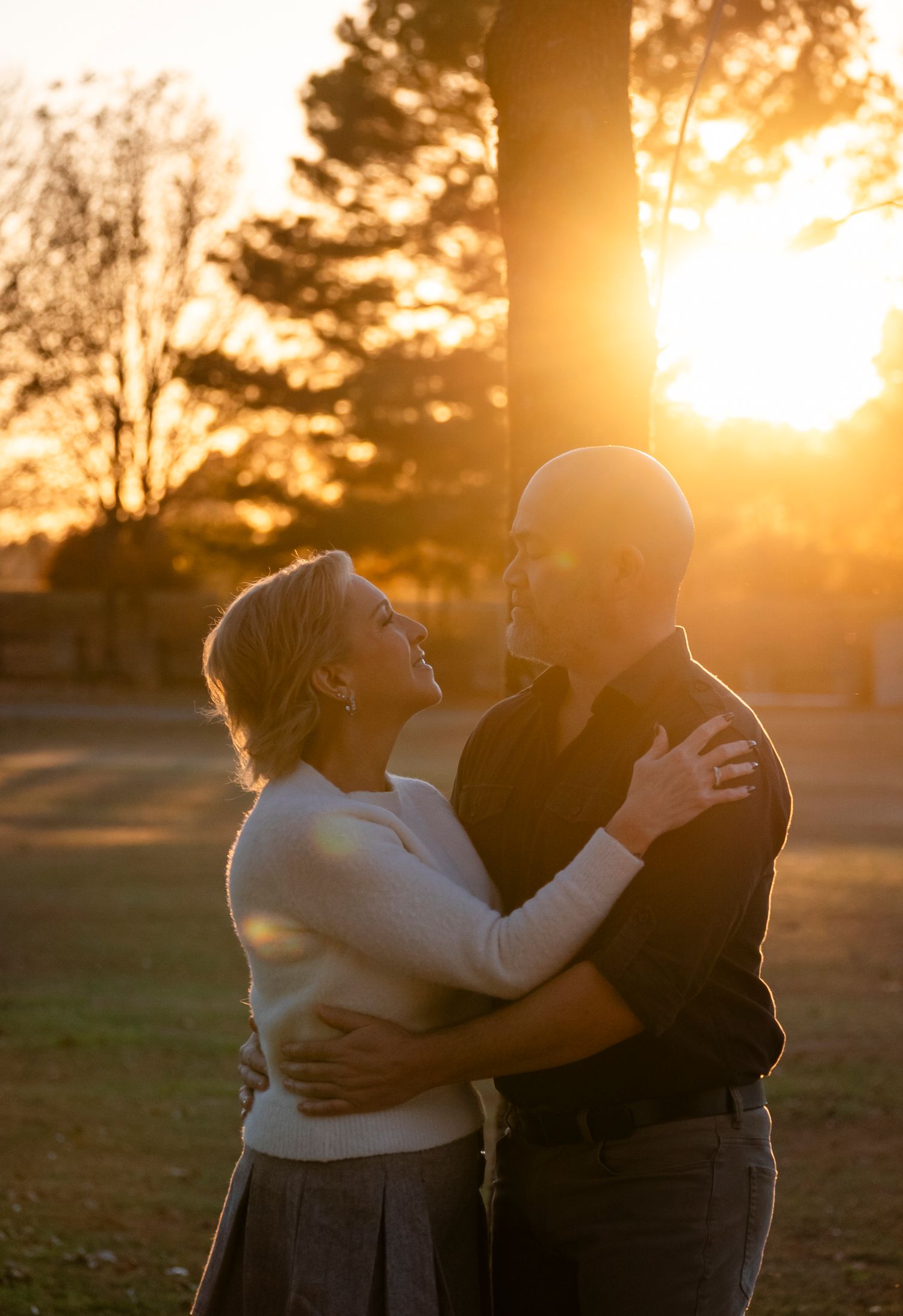 Romantic couple embracing at sunset in a park, warm golden light glowing behind them as they gaze at each other.