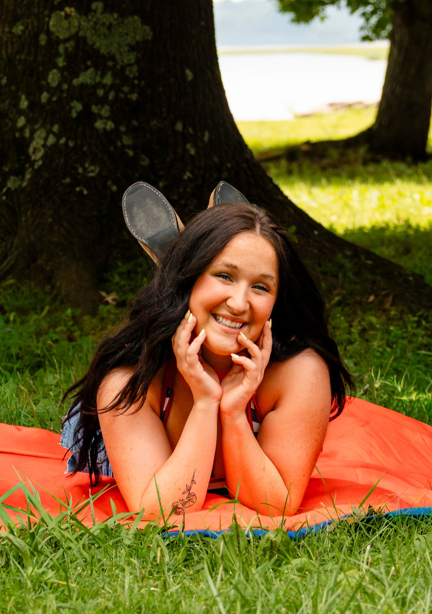 Smiling young woman with bunny ears lying on orange blanket in green park under tree on sunny day.