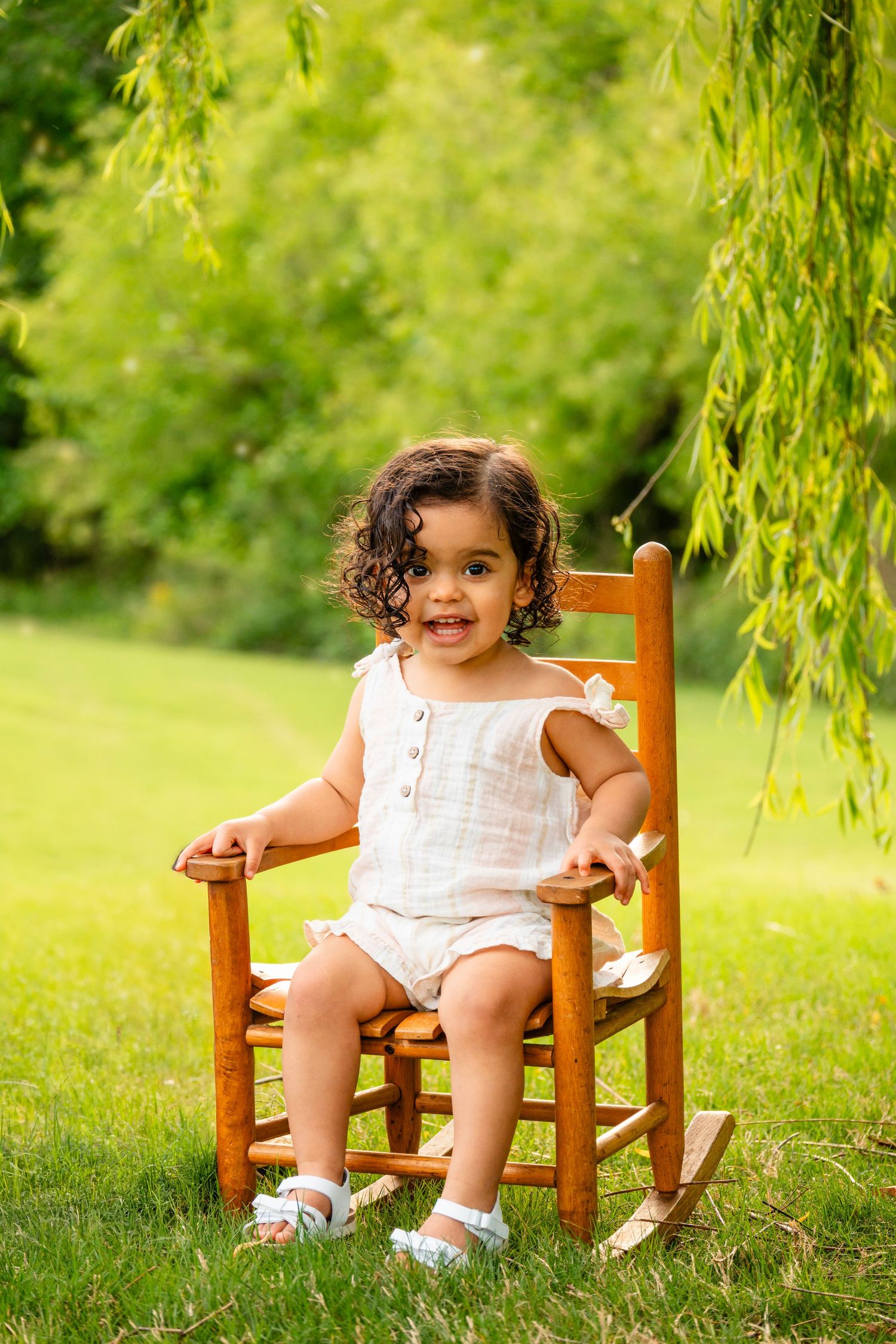 Toddler girl in white romper sitting in a wooden rocking chair outdoors on green grass under a willow tree.