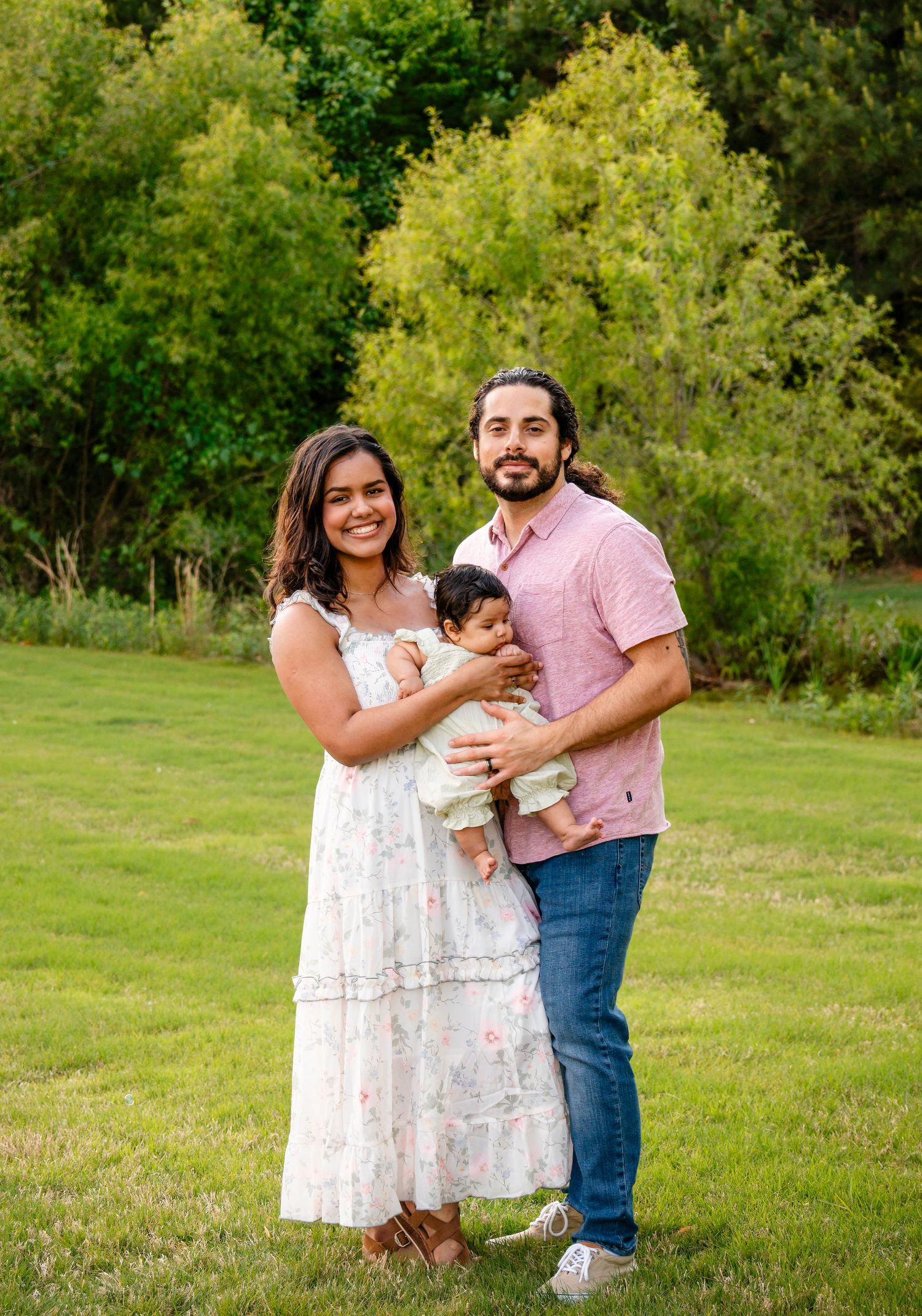 Young family with a baby posing outdoors on green grass with lush trees in the background.
