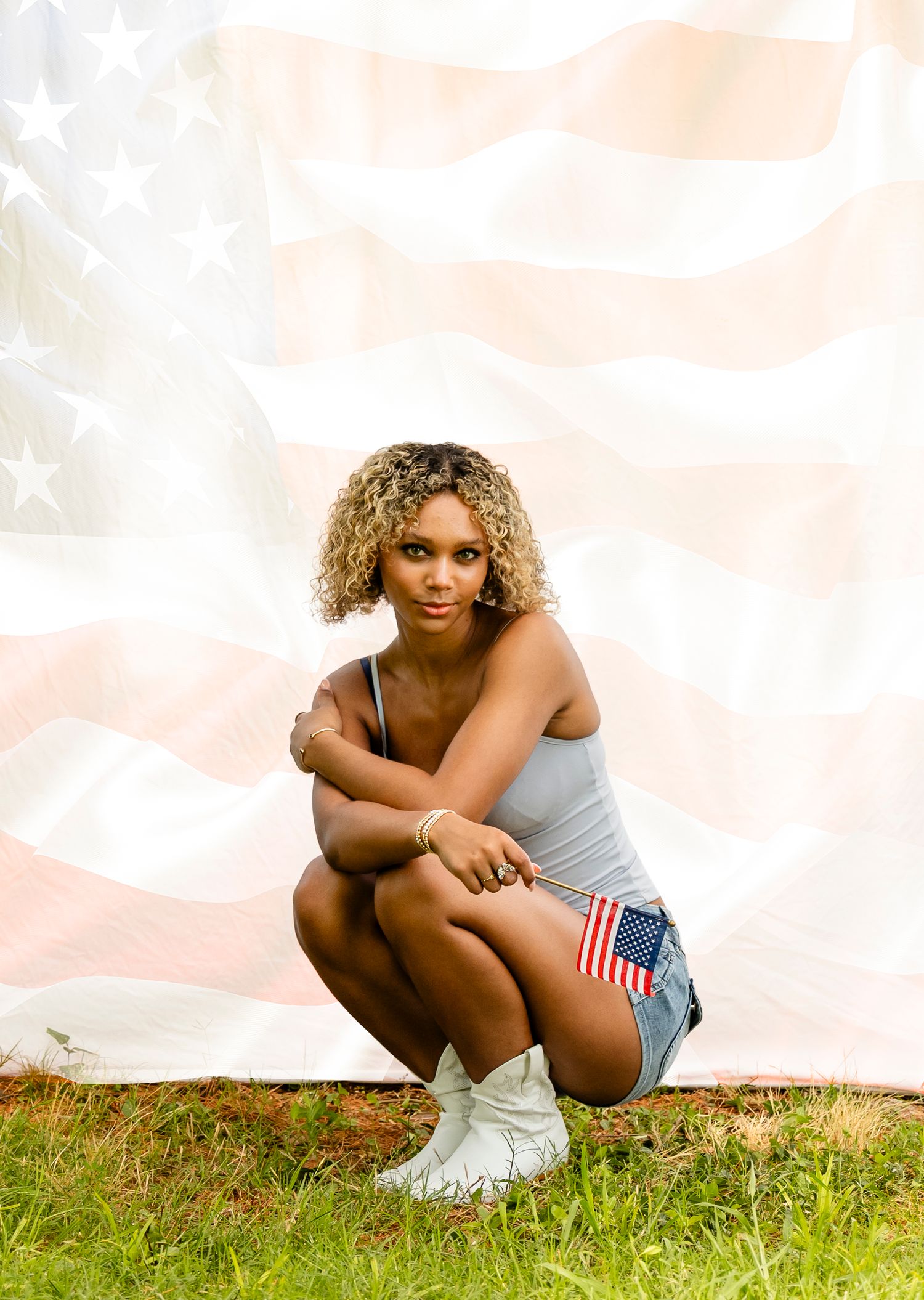 Young woman in denim shorts and white boots crouching in front of a large American flag backdrop.
