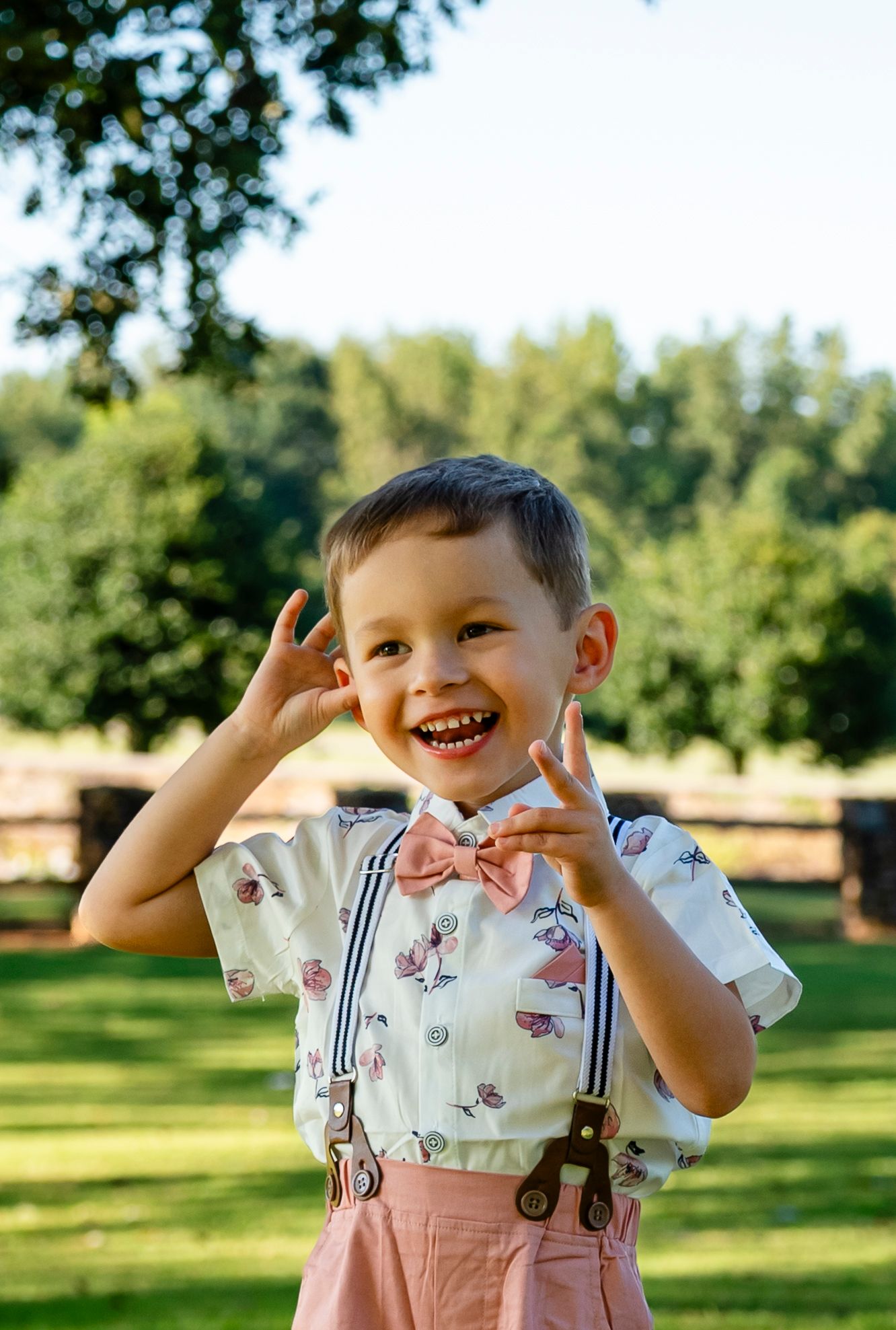 Smiling young boy in pink bow tie, suspenders, and patterned shirt posing outdoors in a sunny green park.