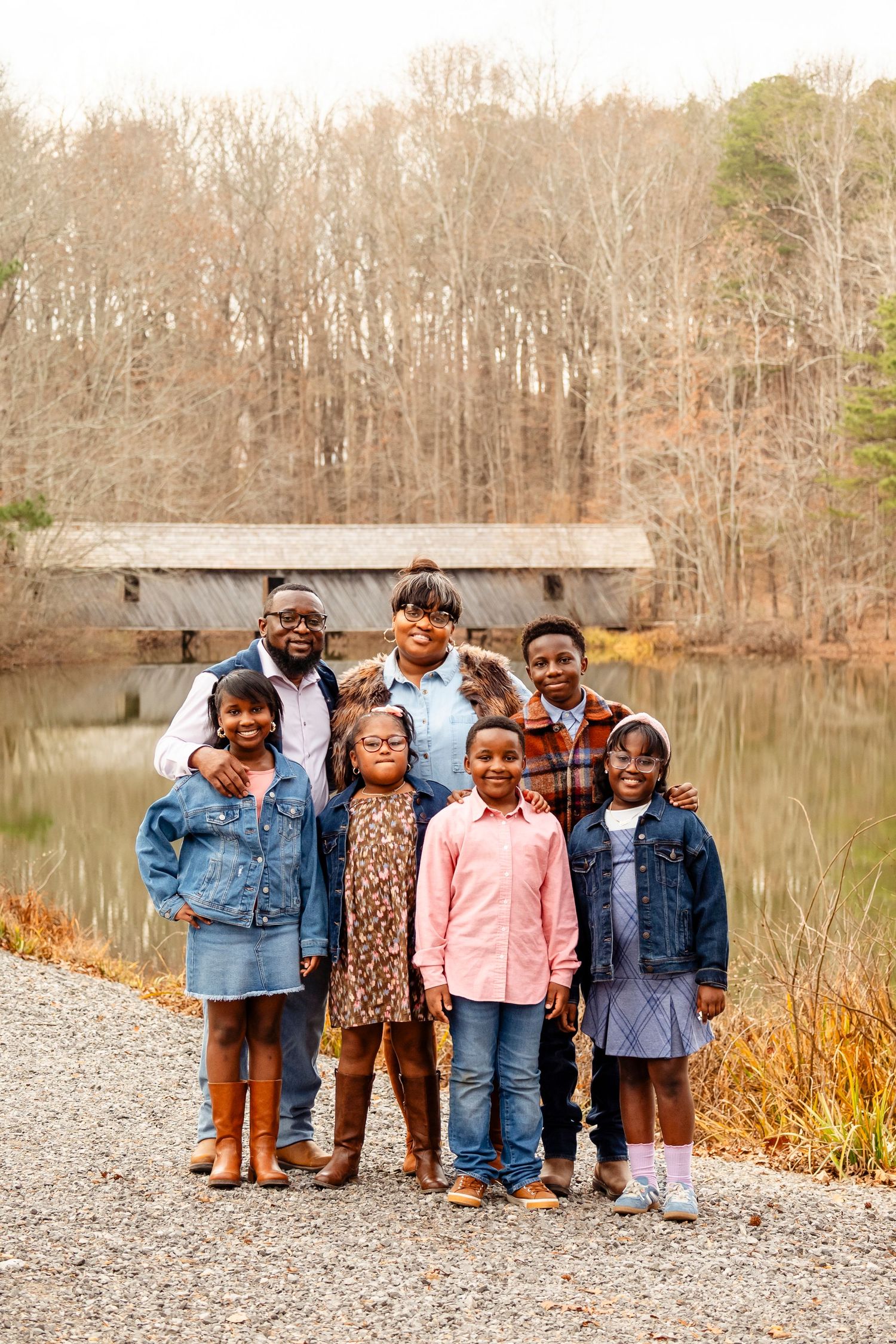 A Black family of seven poses together near a pond with a covered bridge in an autumn setting.