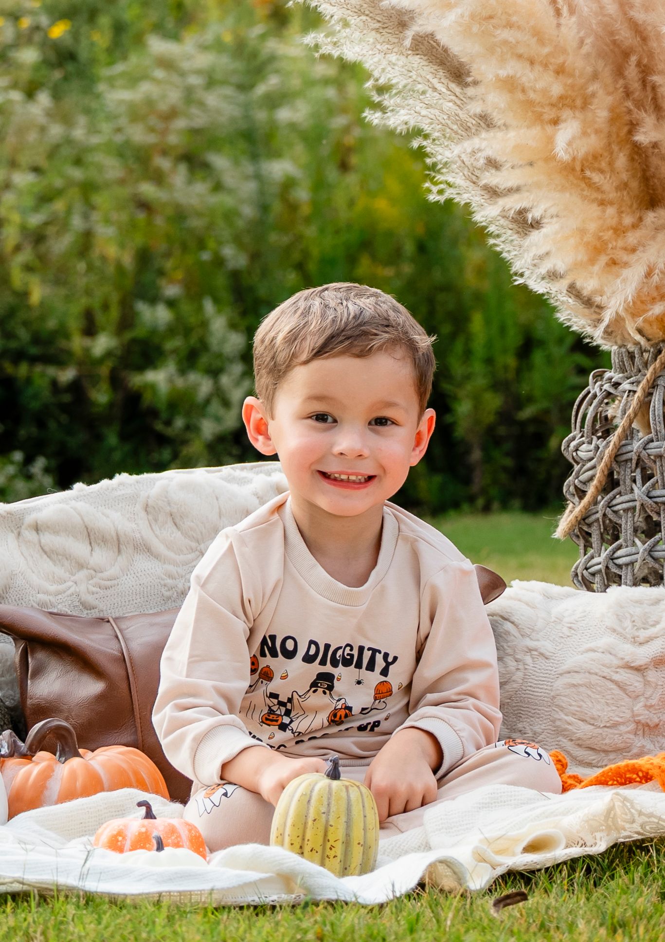 Cheerful boy in beige sweatshirt sitting among pumpkins with fall decorations in garden.