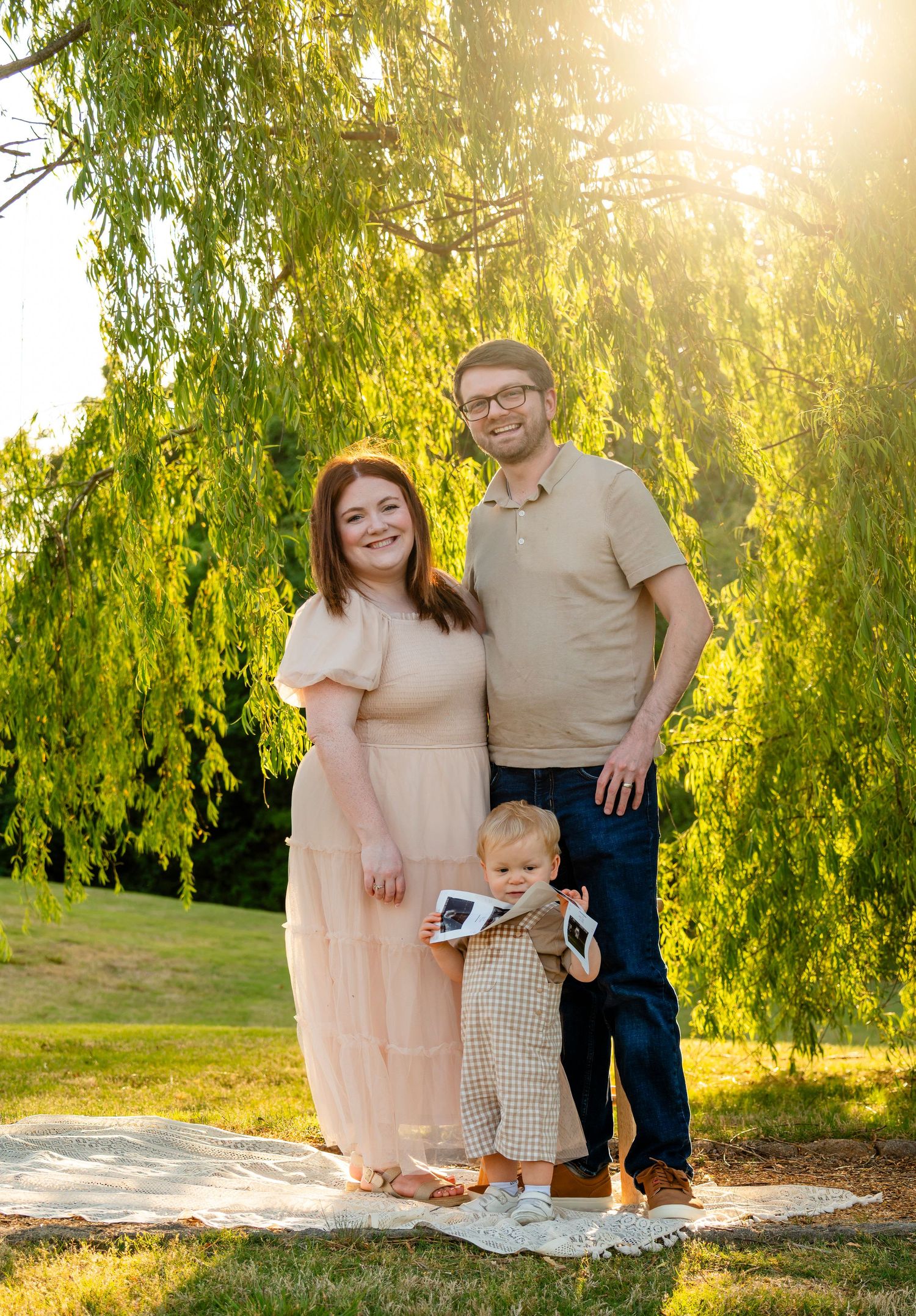 Pregnant woman in pink dress with husband and toddler holding ultrasound under sunlit willow tree.