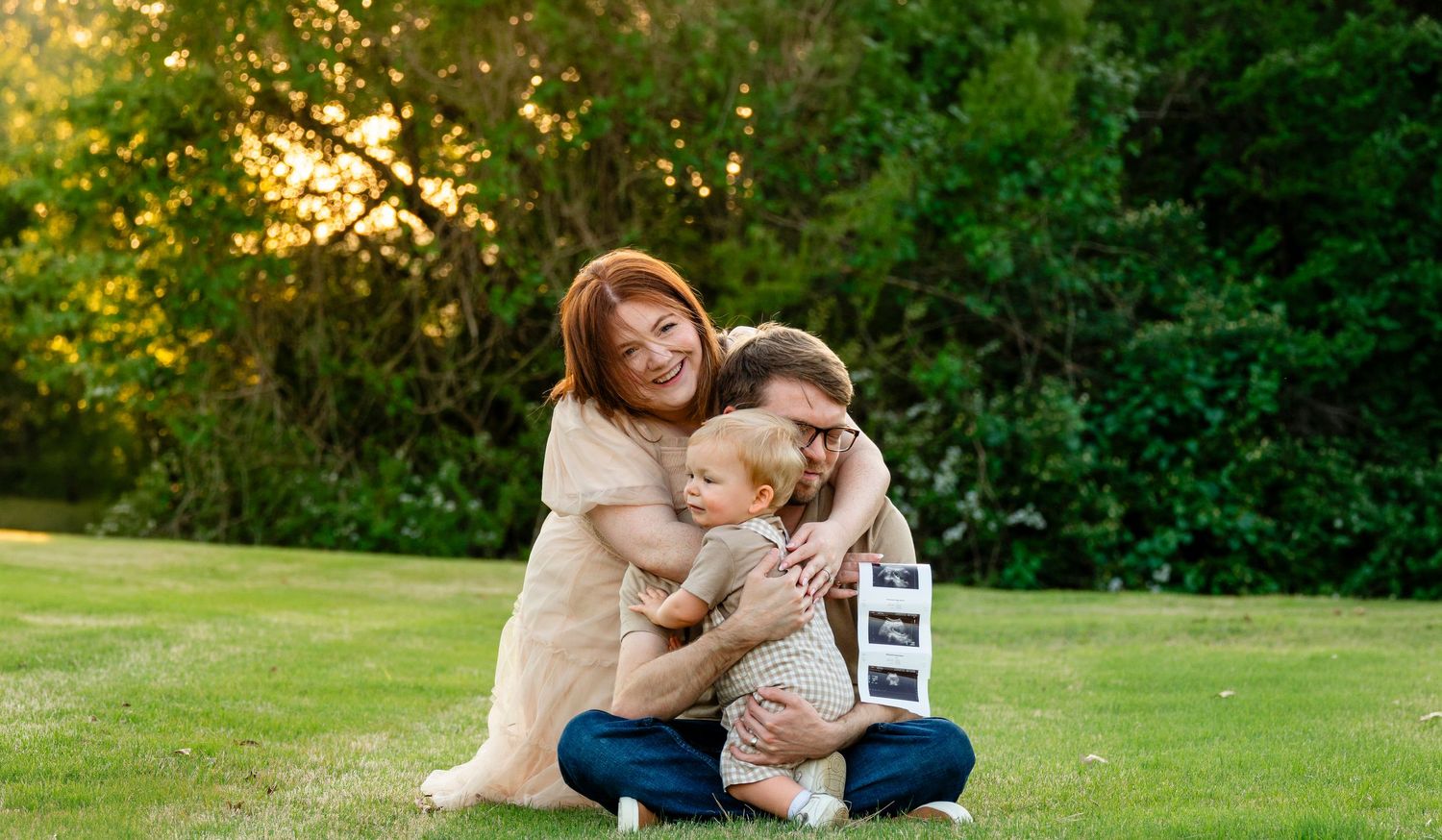 Happy family maternity announcement on grass with parents hugging toddler holding ultrasound photos.