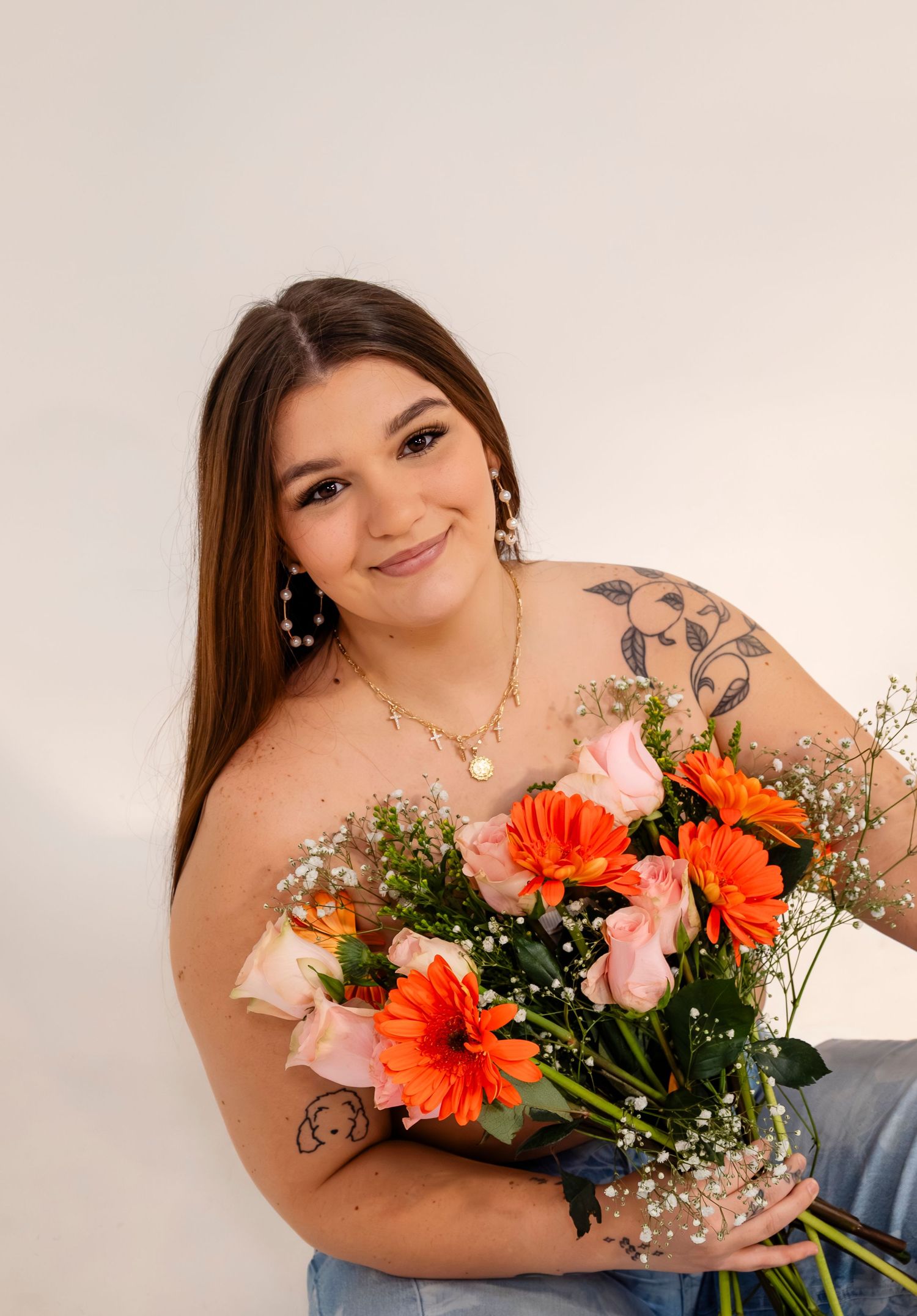 Smiling brunette woman with tattoos holds a vibrant bouquet of orange gerberas and pink roses.