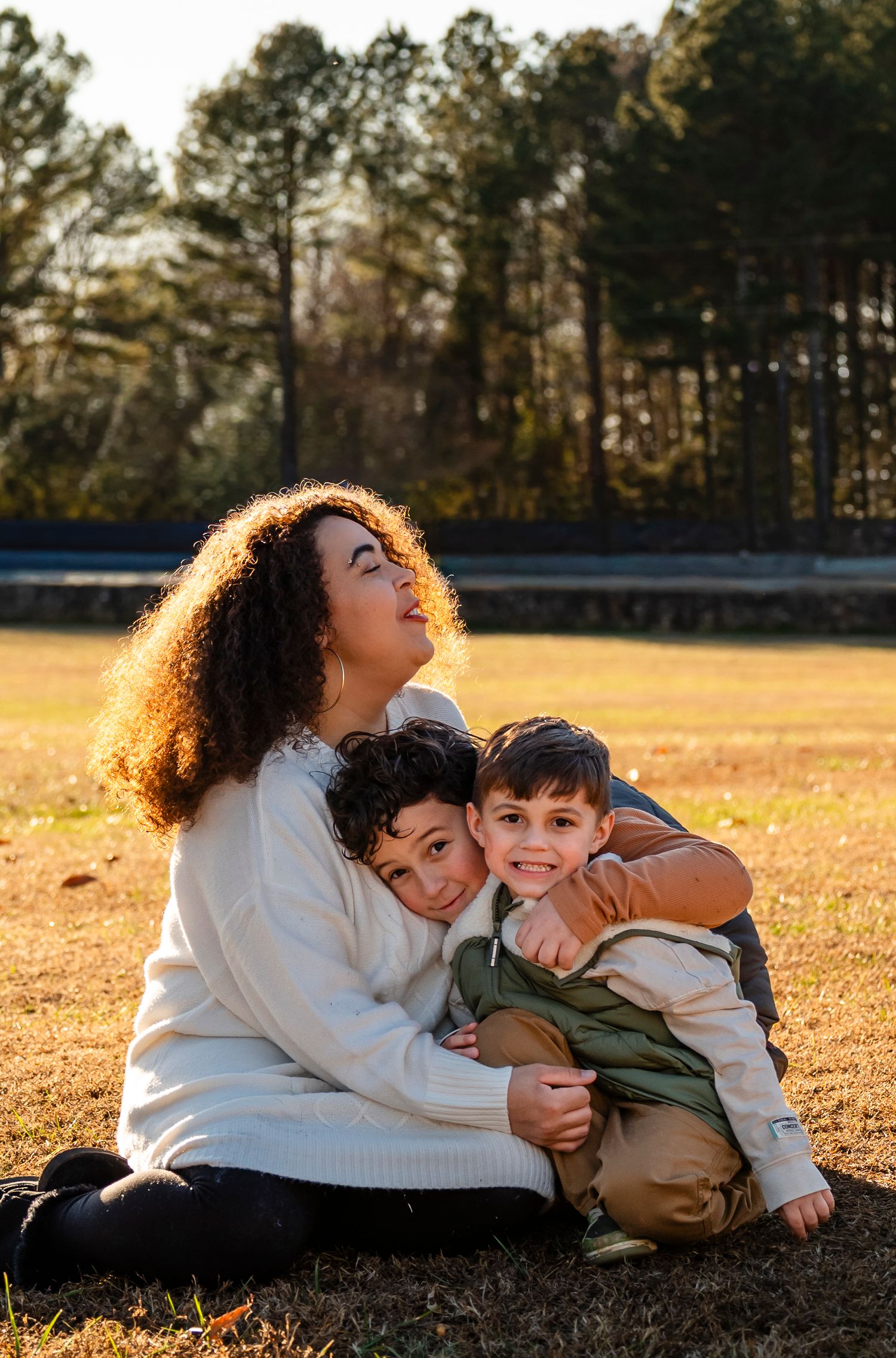 A mother sits on autumn grass hugging her two young sons during a warm golden hour outdoor session.